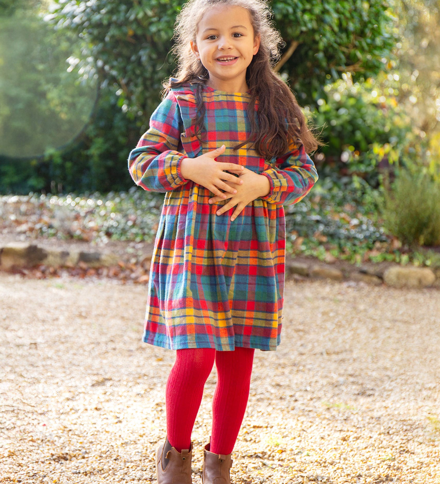 A child stood outside on a lawn with trees in the background, wearing the Frugi Agnes Dress - Merry Rainbow Check