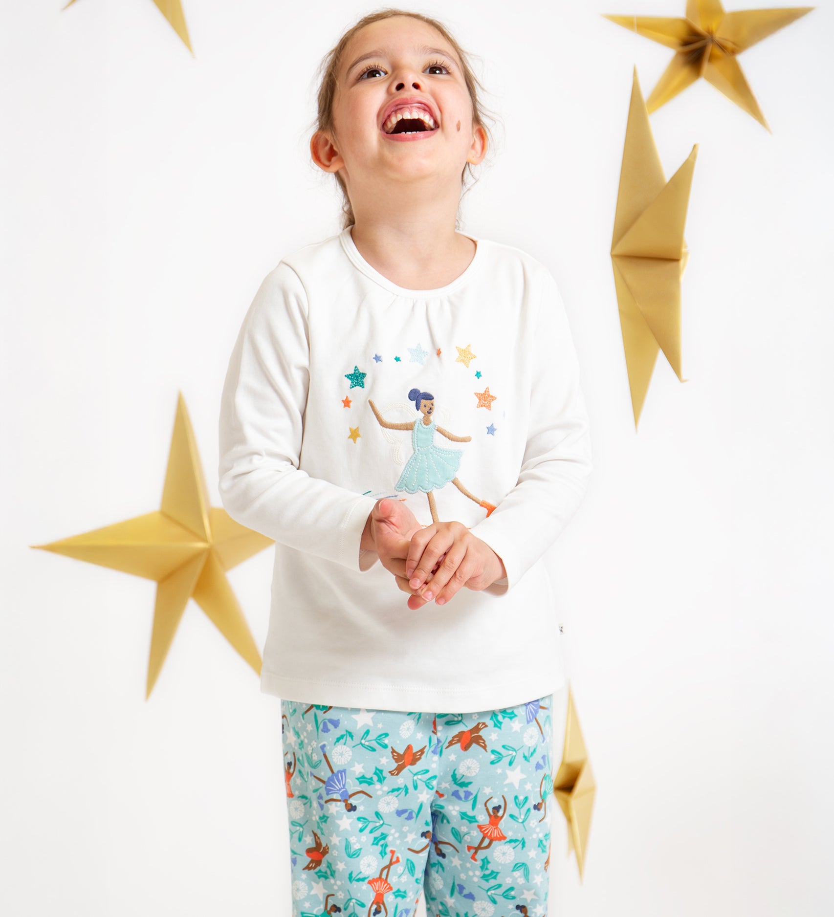 A child looking up and smiling, wearing the Frugi Alana Pyjamas - Glacier Enchanted Forest/Skater. With a white background and golden stars