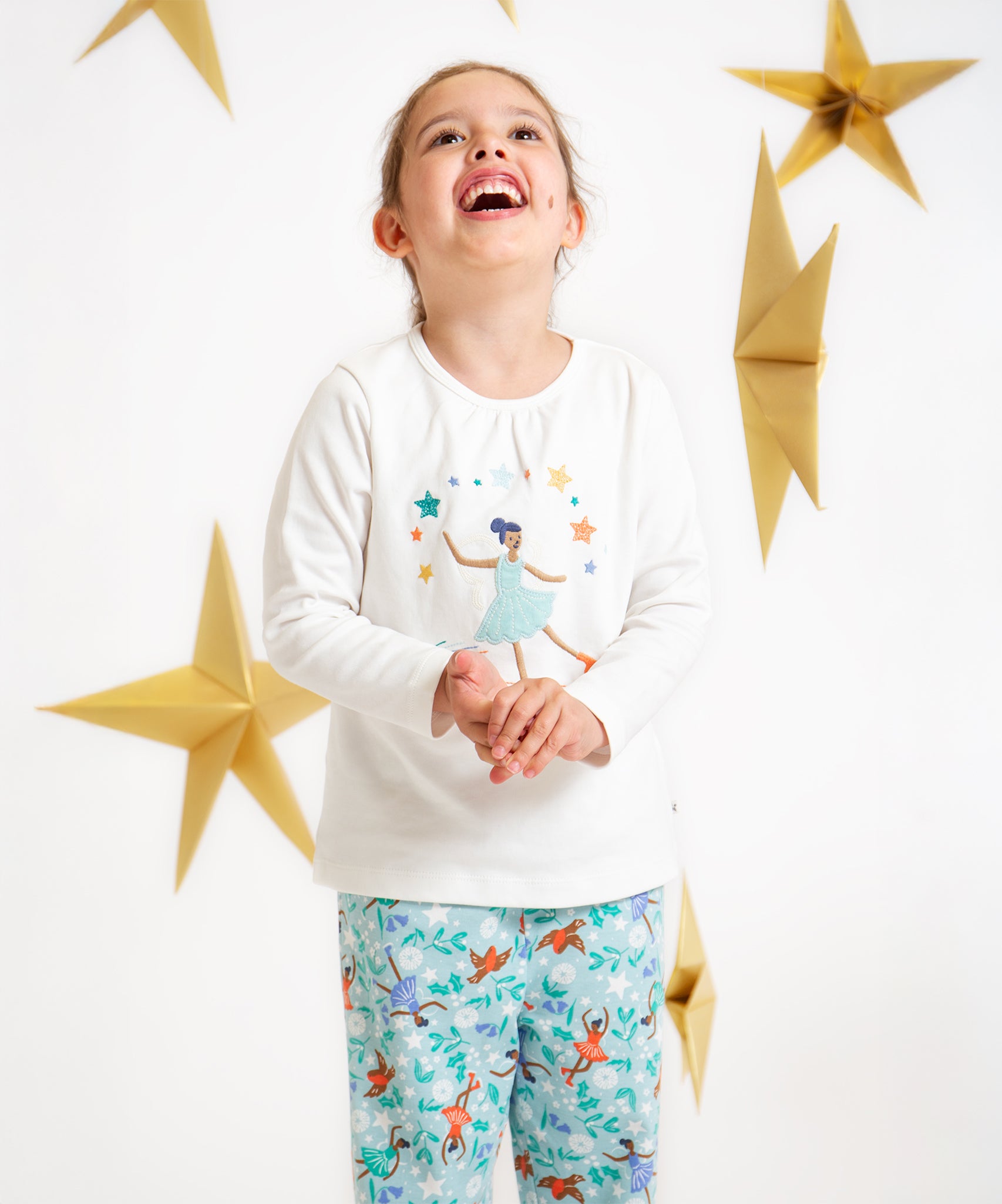 A child looking up and smiling, wearing the Frugi Alana Pyjamas - Glacier Enchanted Forest/Skater. With a white background and golden stars