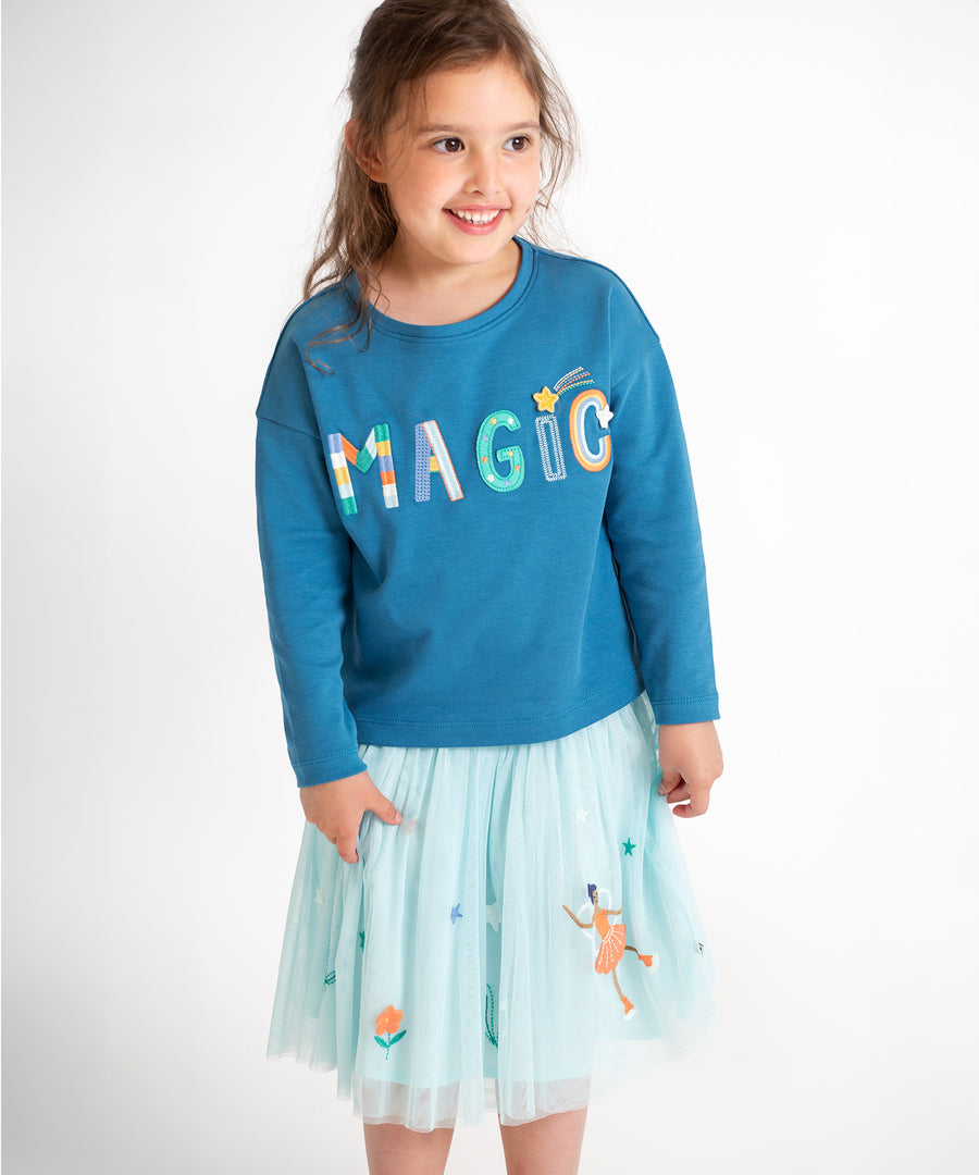 A child stood up and smiling, wearing the Frugi Scarlet Twirly Skirt - Glacier/Fairy, and a blue Frugi top that says "Magic" across the front