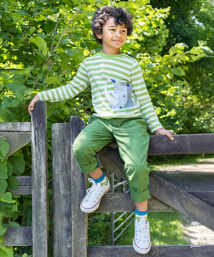 A child sat on a wooden gate and fence post, wearing the Frugi Pioneer Trousers - Jungle Green