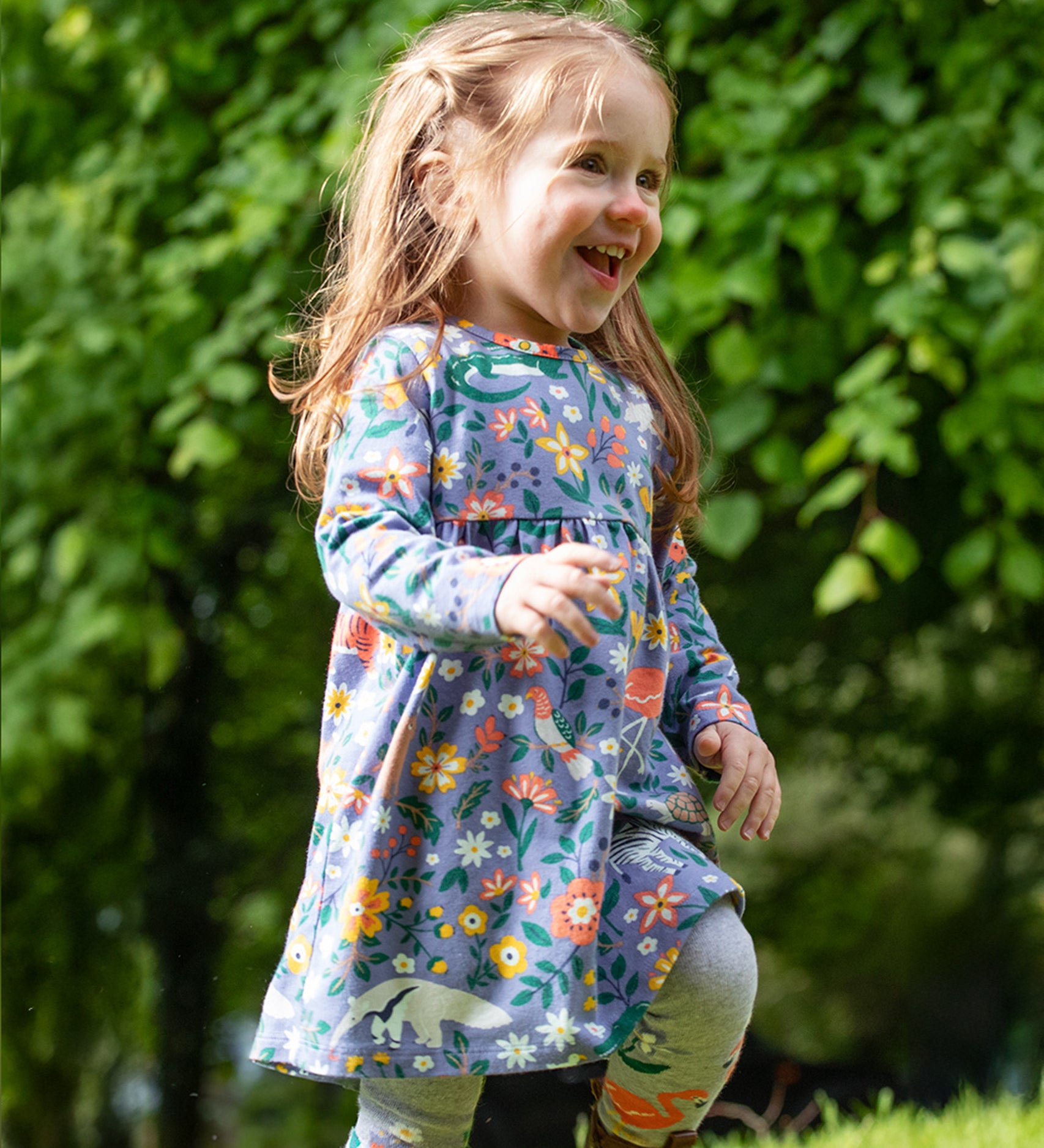 A child, happily running outside on grass in front of a tree line, is wearing the Frugi Dee Dress - Rainforest Friends. The dress is a lovely lilac colour, with various rainforest animals and flowers all over