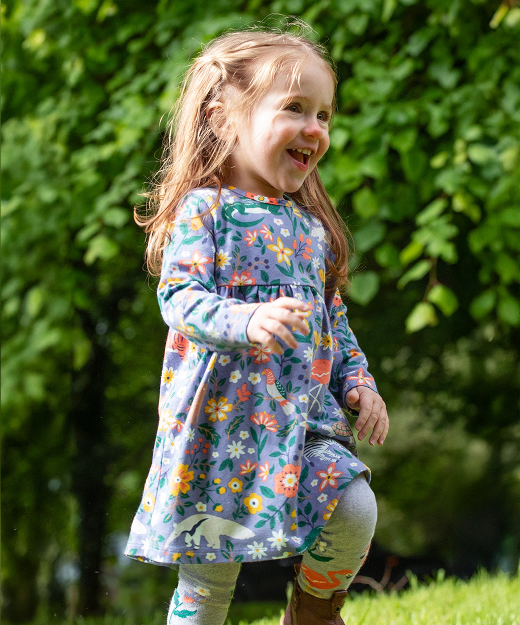 A child, happily running outside on grass in front of a tree line, is wearing the Frugi Dee Dress - Rainforest Friends. The dress is a lovely lilac colour, with various rainforest animals and flowers all over