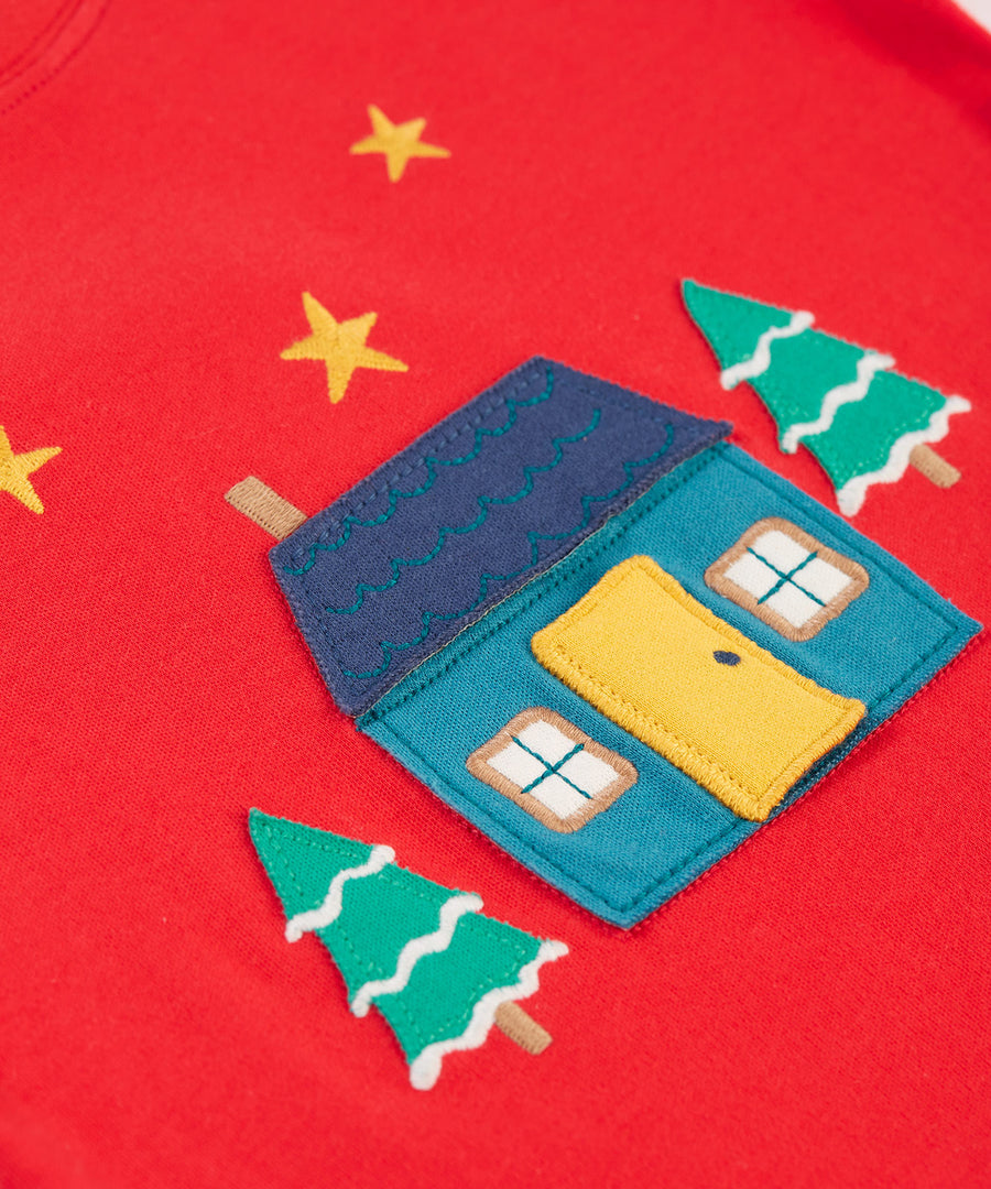 A closer look at the home applique on the Frugi Ira Interactive Applique Top - True Red/Winter Cottage. The house is blue with a navy blue roof, a yellow door and the snowy trees sit either side of the house