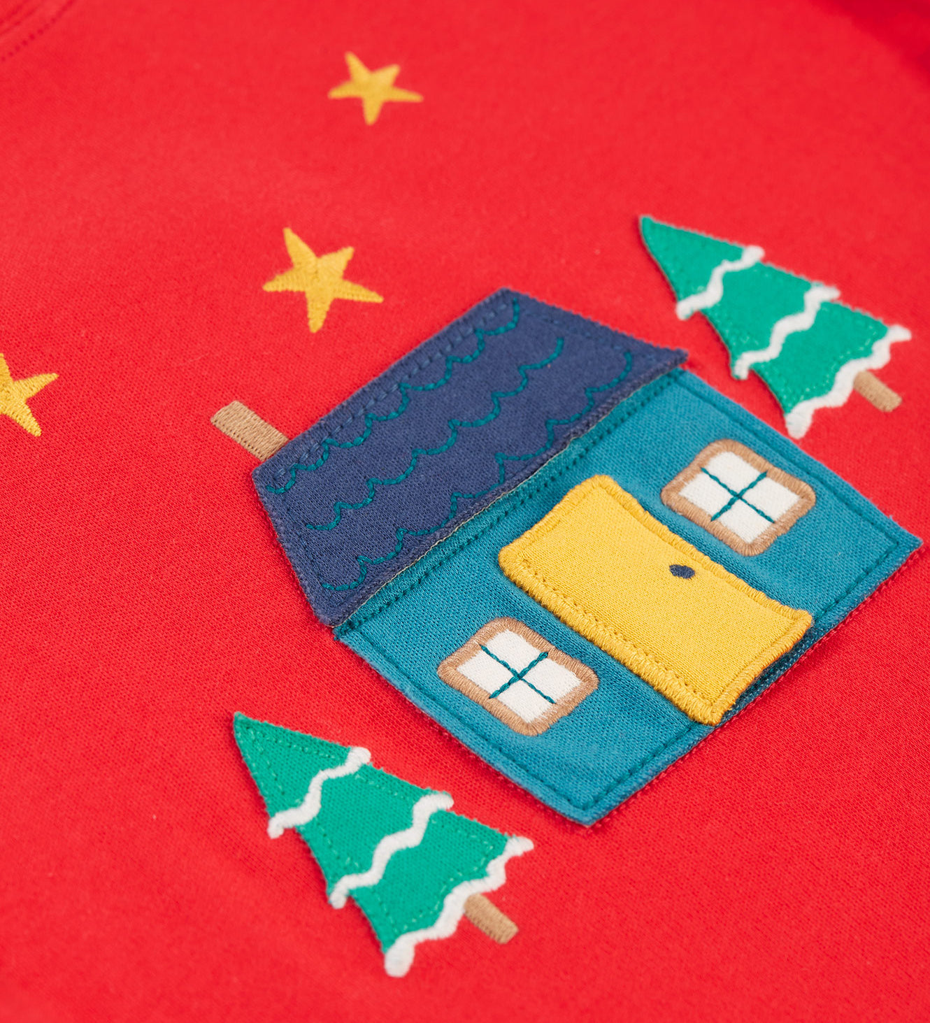 A closer look at the home applique on the Frugi Ira Interactive Applique Top - True Red/Winter Cottage. The house is blue with a navy blue roof, a yellow door and the snowy trees sit either side of the house