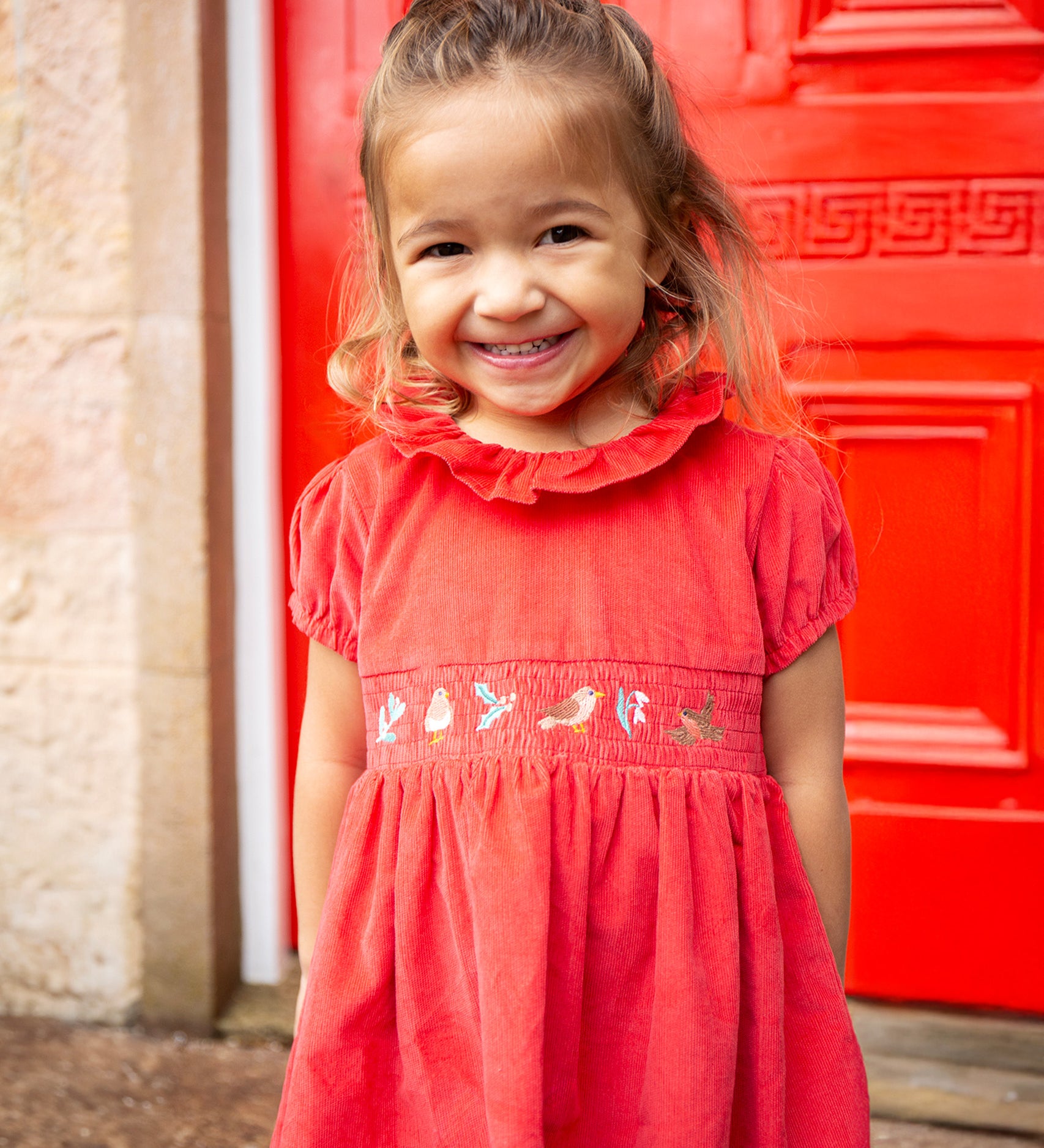 A child stood in front of a red door, wearing the Frugi Amilie Party Dress - True Red/Robin