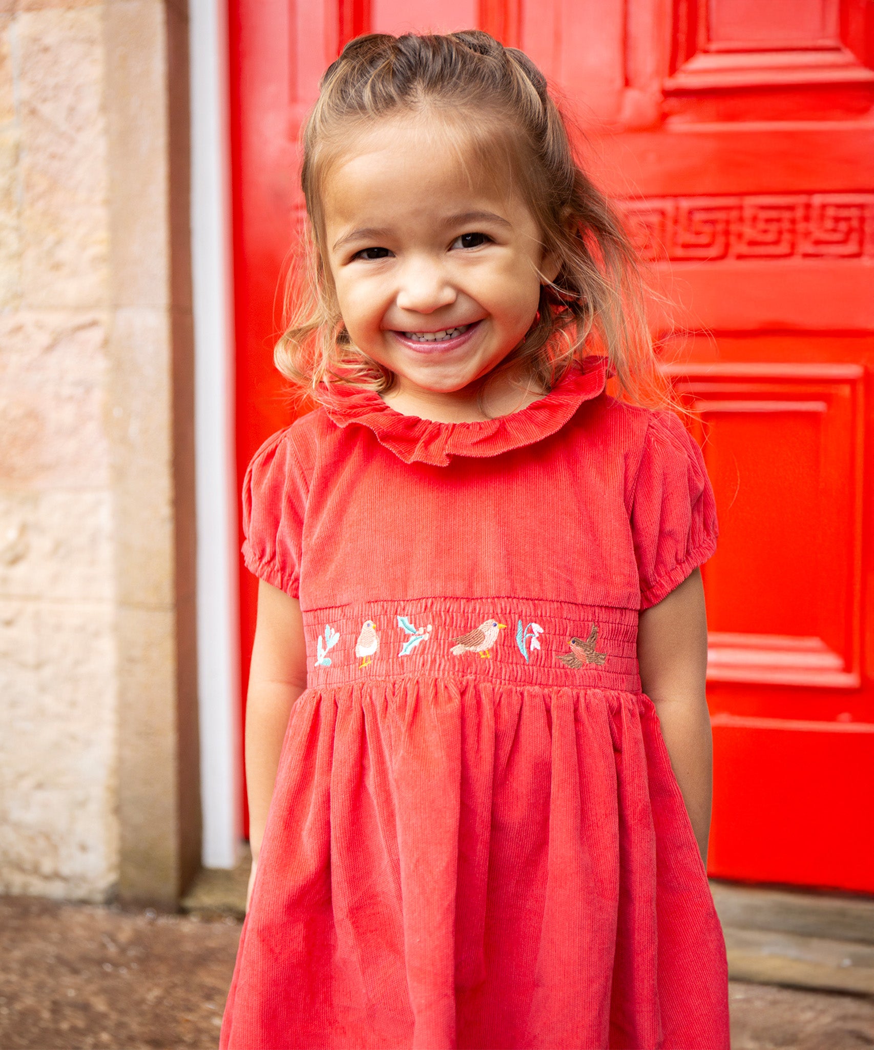 A child stood in front of a red door, wearing the Frugi Amilie Party Dress - True Red/Robin