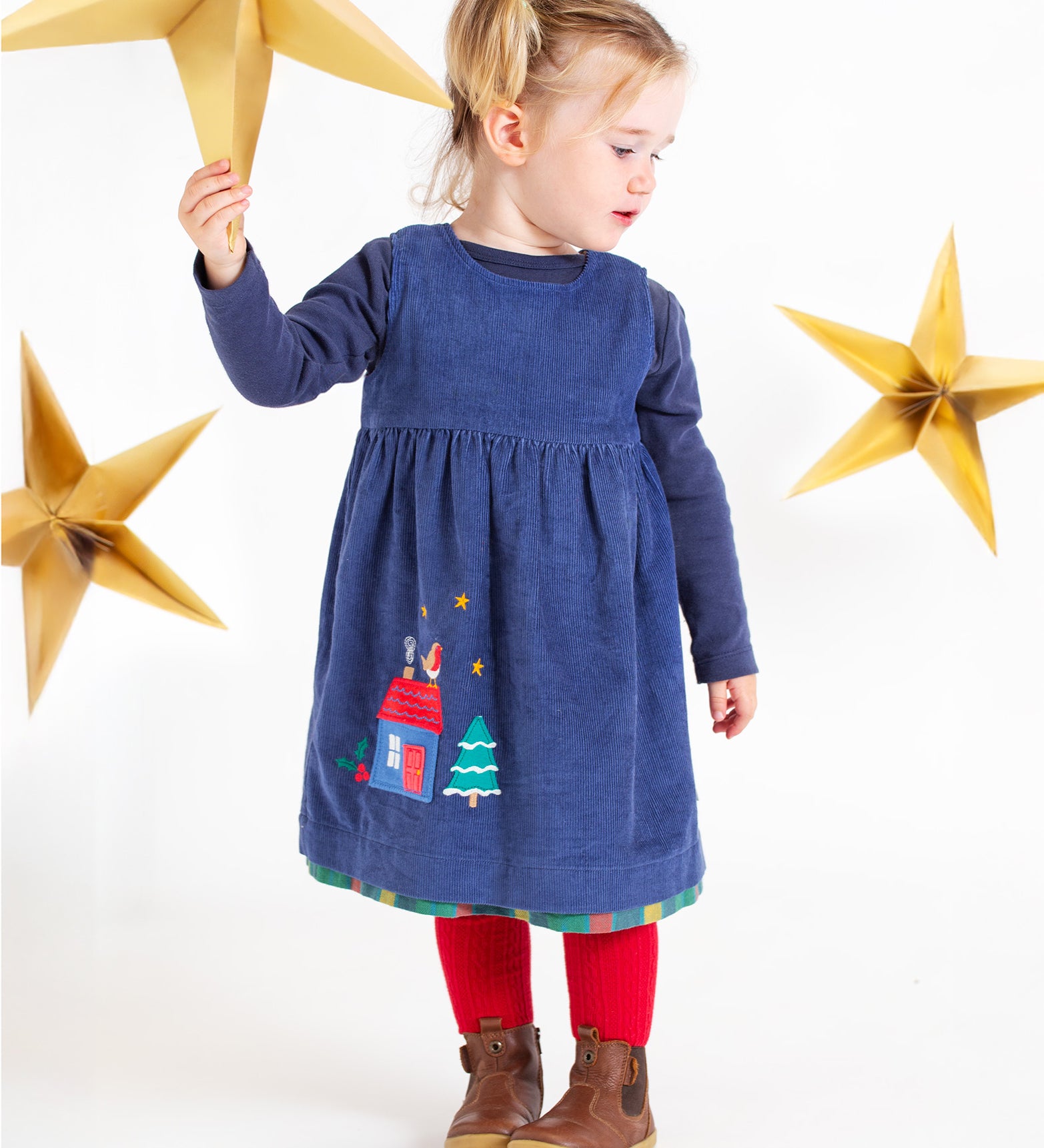 A child holding a gold paper star, with gold stars in the background. The child is wearing the Frugi Inara Cord Reversible Dress - Navy Blue/Rainbow Check, with the blue corduroy side showing