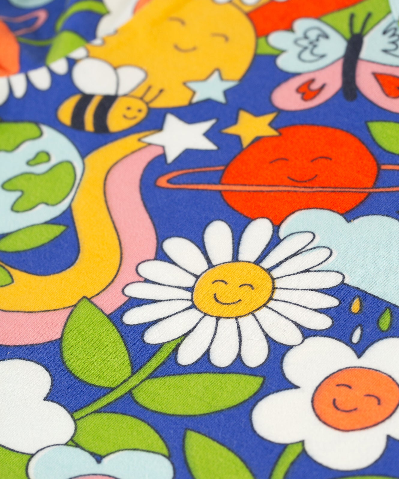 The Retro Happy print on the Frugi Sofia Skater Dress - Retro Happy. The image shows smiling planets, flowers, bees, rainbow swirls, stars and butterflies on a royal blue fabric