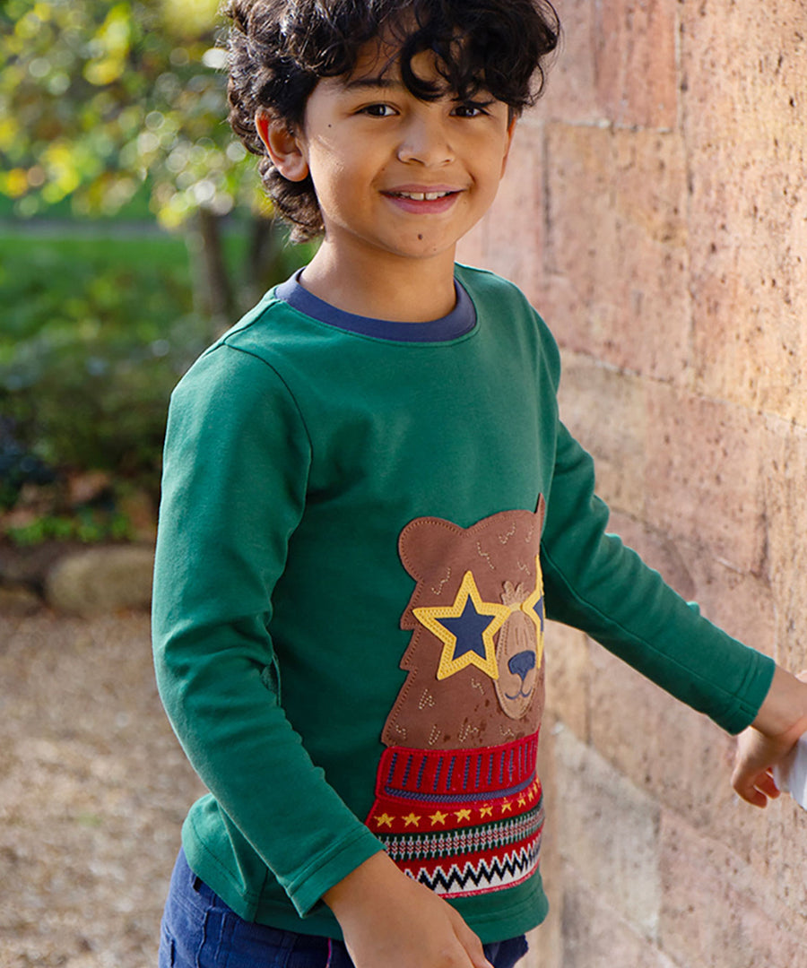 A child, stood outside next to a large brick wall, wearing the Frugi Terrific Applique Top - Teal/Fairisle Bear