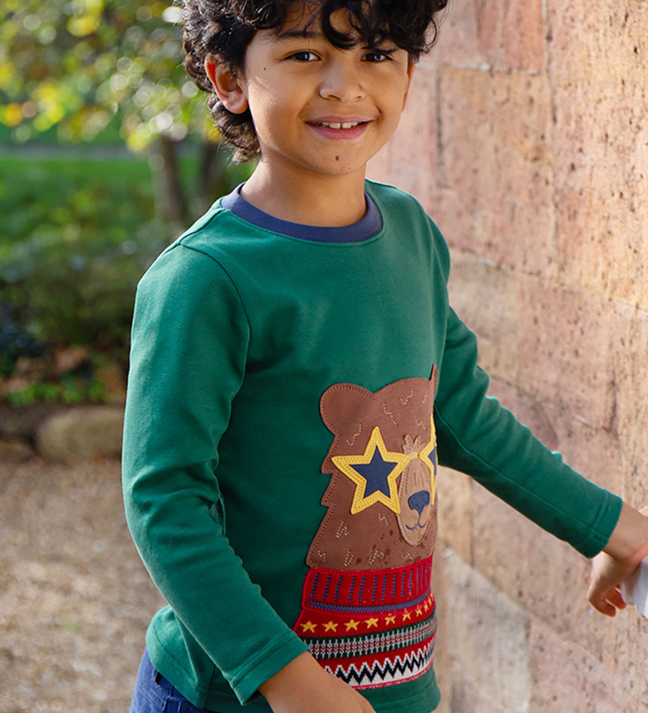 A child, stood outside next to a large brick wall, wearing the Frugi Terrific Applique Top - Teal/Fairisle Bear