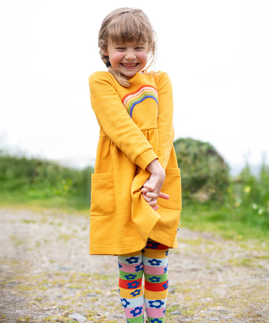 A child happily wearing the Frugi Niamh Dress - Gold/Rainbow. The dress is paired with the Frugi Retro Happy Norah Tights