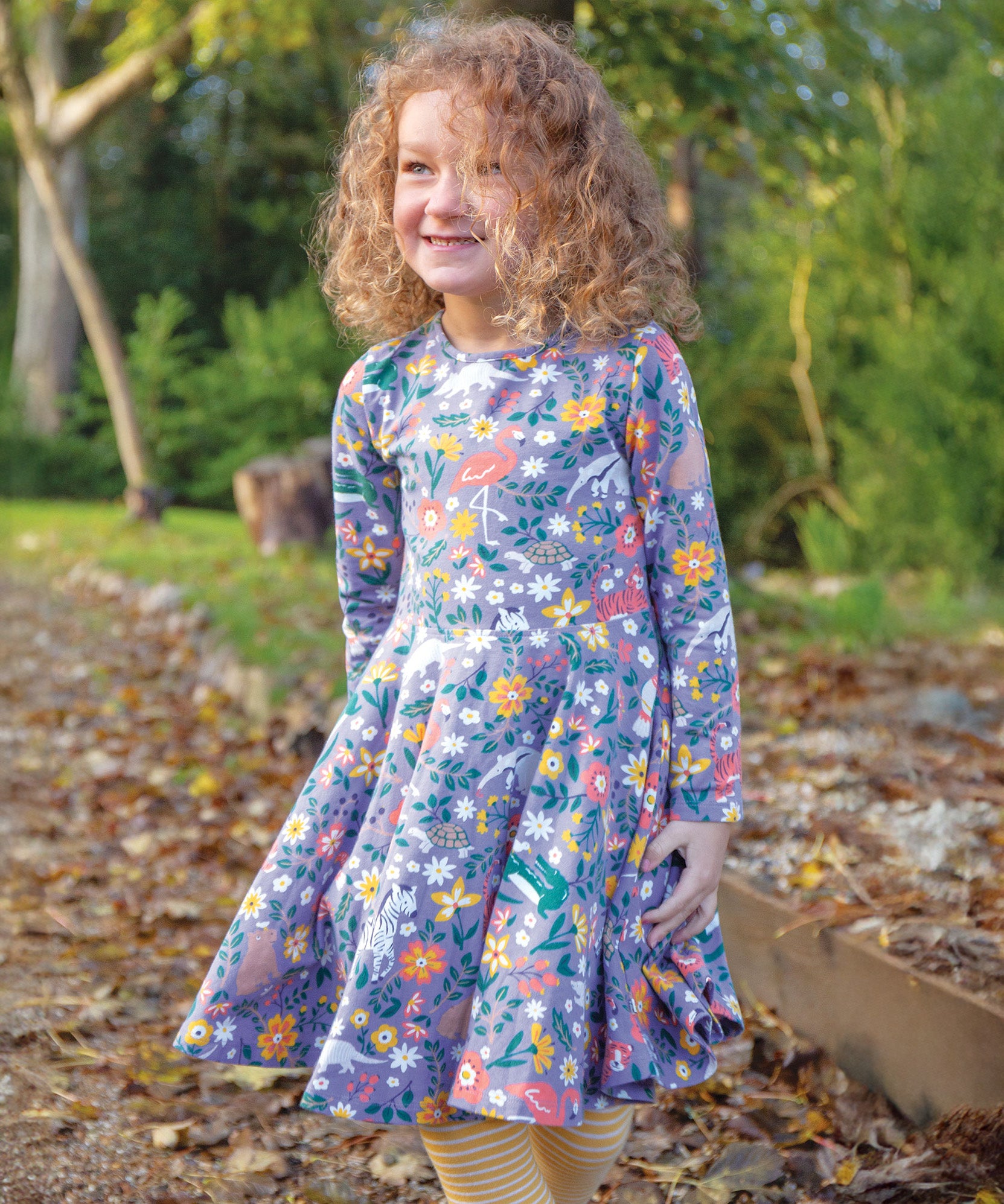 A child happily stood outside on a path with crunchy autumn leaves, wearing the Frugi Sofia Skater Dress - Rainforest Friends