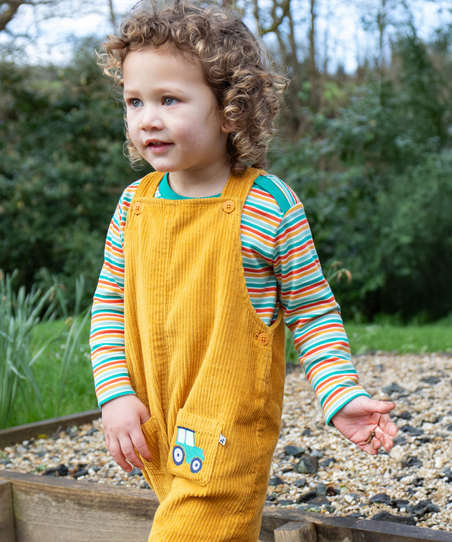 A child outside, wearing the Frugi Adley Cord Dungarees - Gold/Tractor