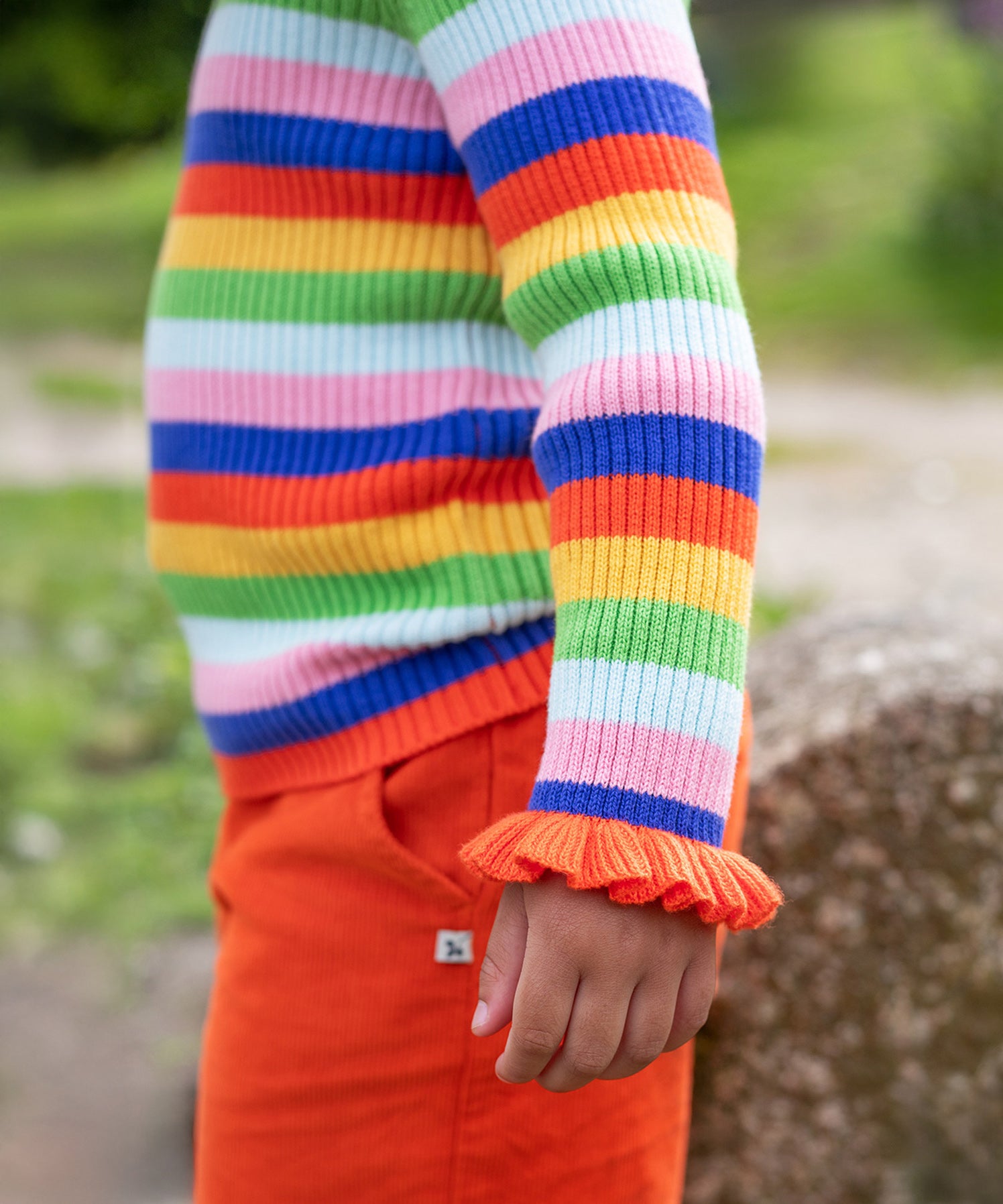 A child is stood outside, wearing the Frugi Zoe Knitted Jumper - Retro Stripe. The image shows a close up of the child's arm and the ruffled wrist cuffs cover over the top of the hand. The colour-block stripes look very vibrant n the natural day light