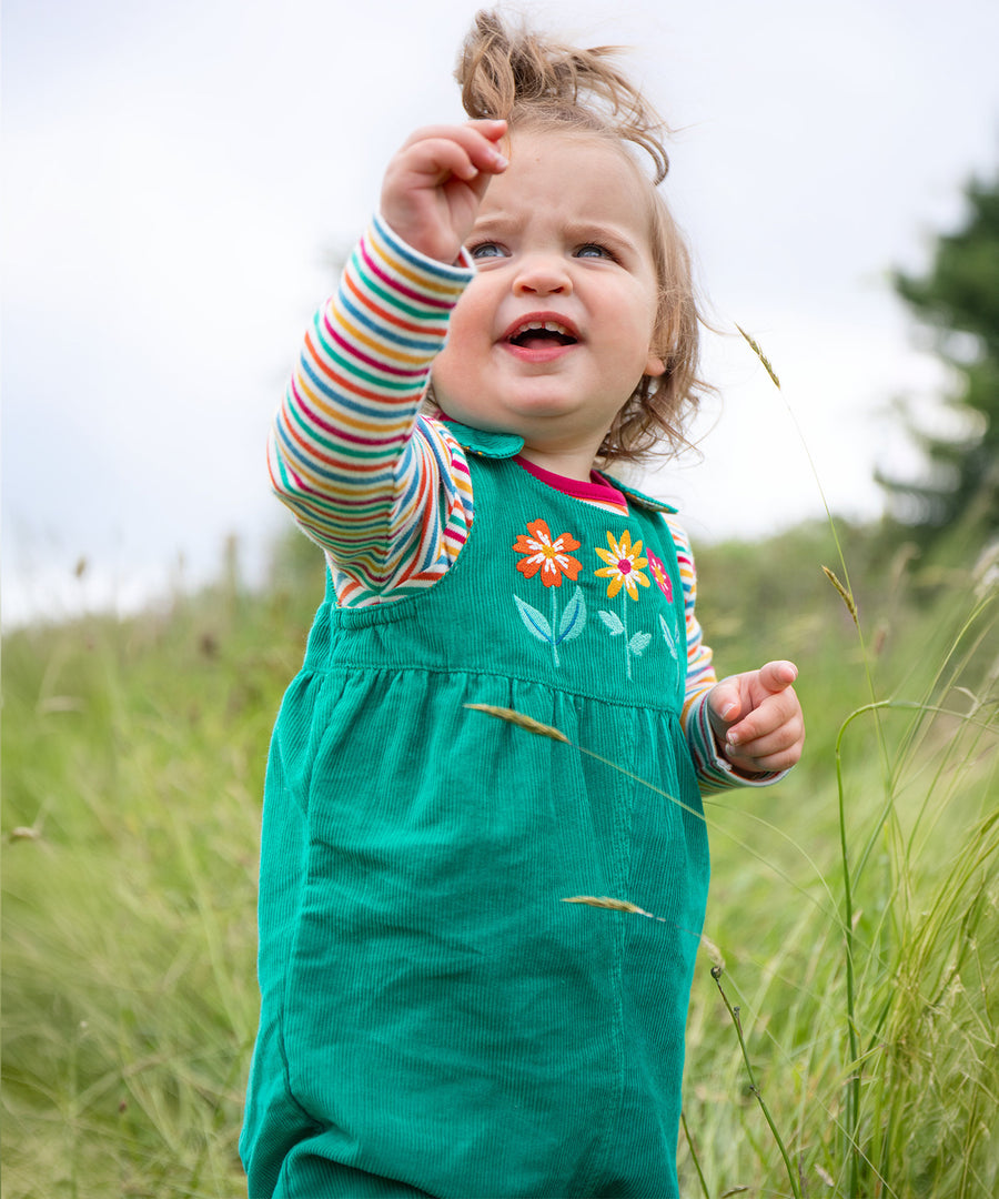 A child playing outside in a grassy field, wearing the Frugi Lacey Reversible Dungaree - Iguana/Autumn Friends