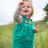 A child playing outside in a grassy field, wearing the Frugi Lacey Reversible Dungaree - Iguana/Autumn Friends