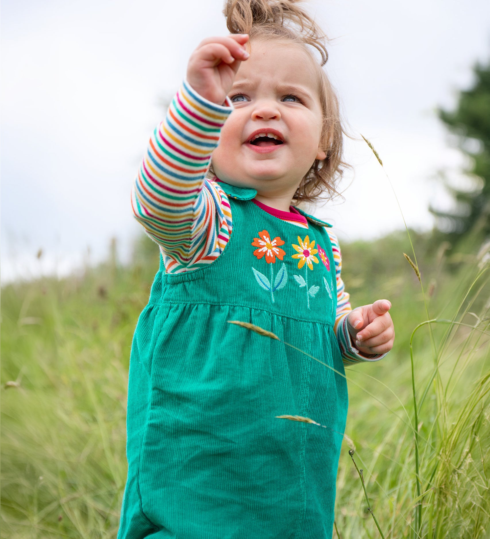 A child playing outside in a grassy field, wearing the Frugi Lacey Reversible Dungaree - Iguana/Autumn Friends