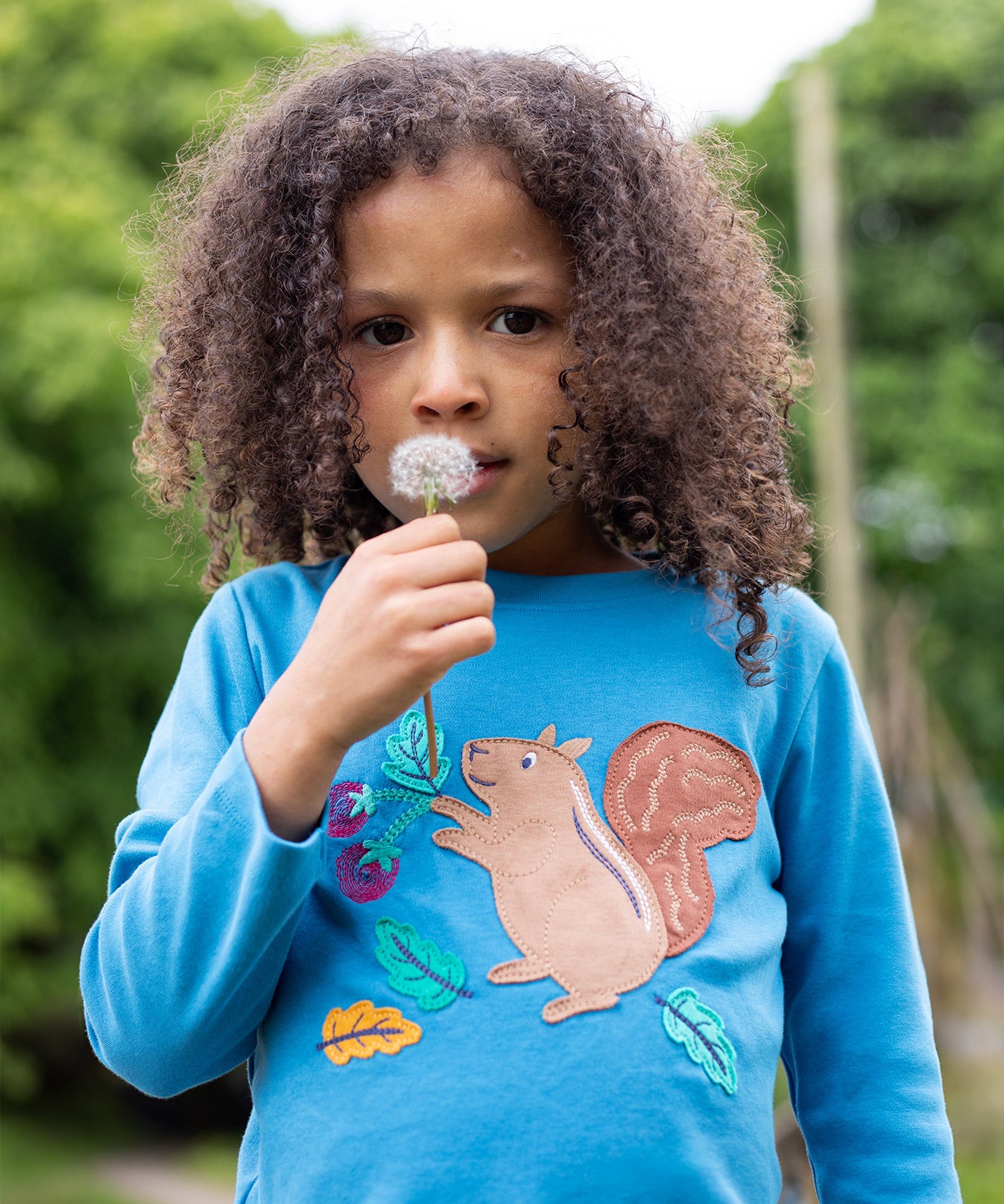 A child blowing a dandelion, whilst wearing the Frugi Adventure Applique Top - Deep Water/Squirrel