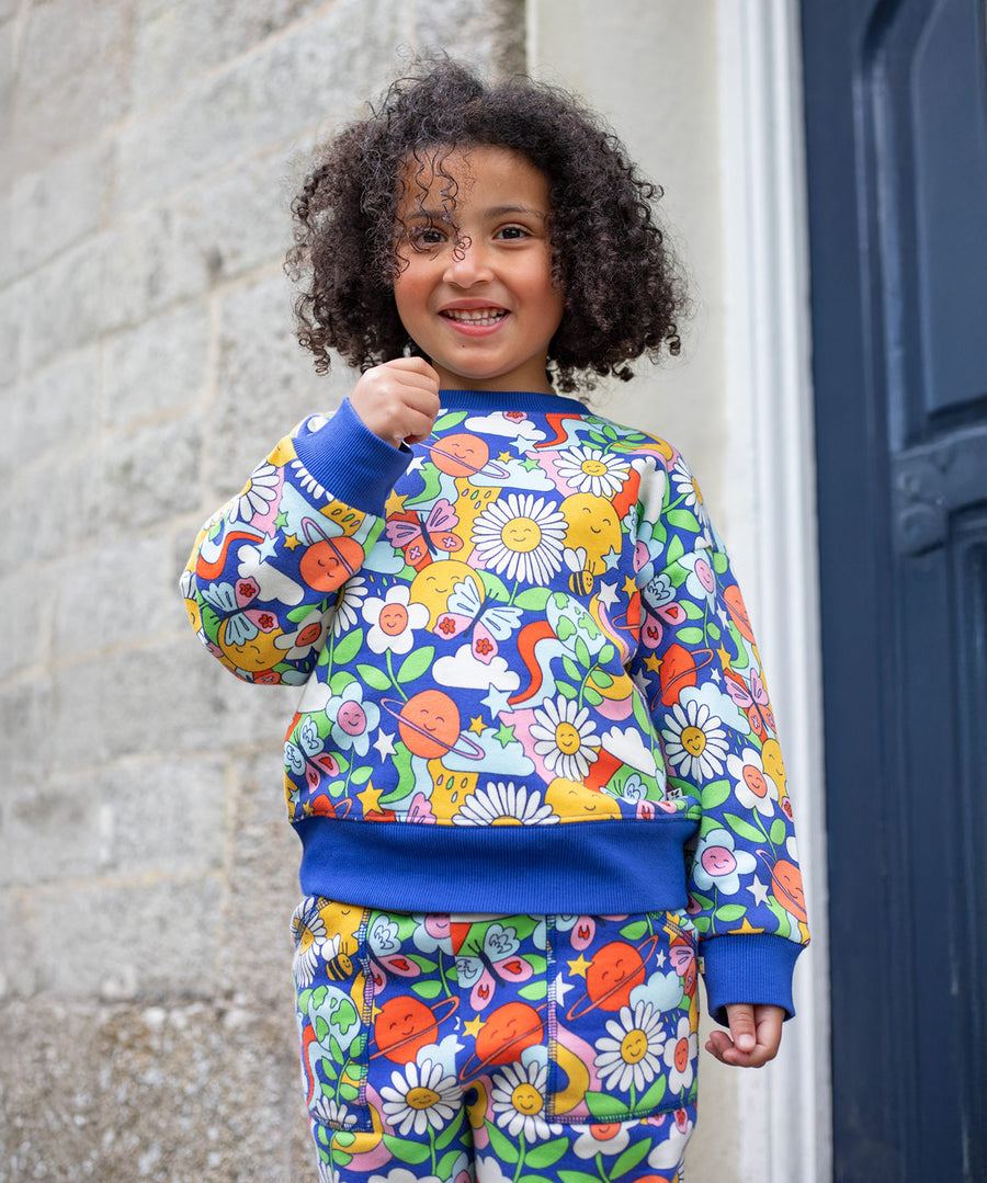 A child outside, stood next to a building with a blue door, is wearing the Frugi Superb Sweatshirt - Retro Happy, and Frugi Retro Happy jogging bottoms