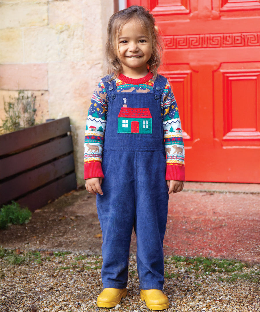 A child smiling and stood in front of a building with a red door, wearing the Frugi Durgan Cord Dungarees - Navy Blue/Winter Cottage.