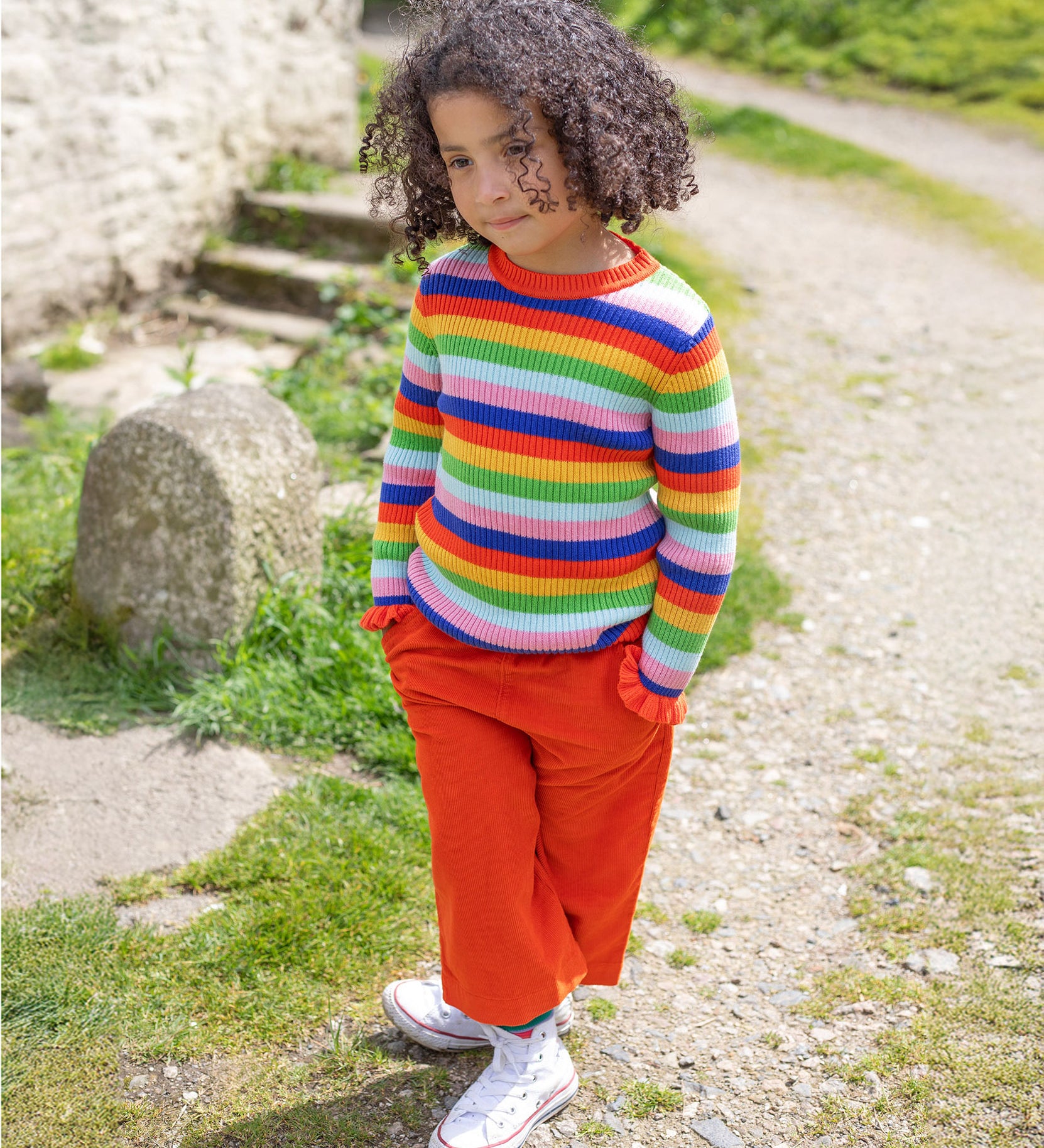 A child is stood outside in the sunshine, with their hands in their pockets. The child wears the Frugi Zoe Knitted Jumper - Retro Stripe, which looks very vibrant in colour in the natural day light