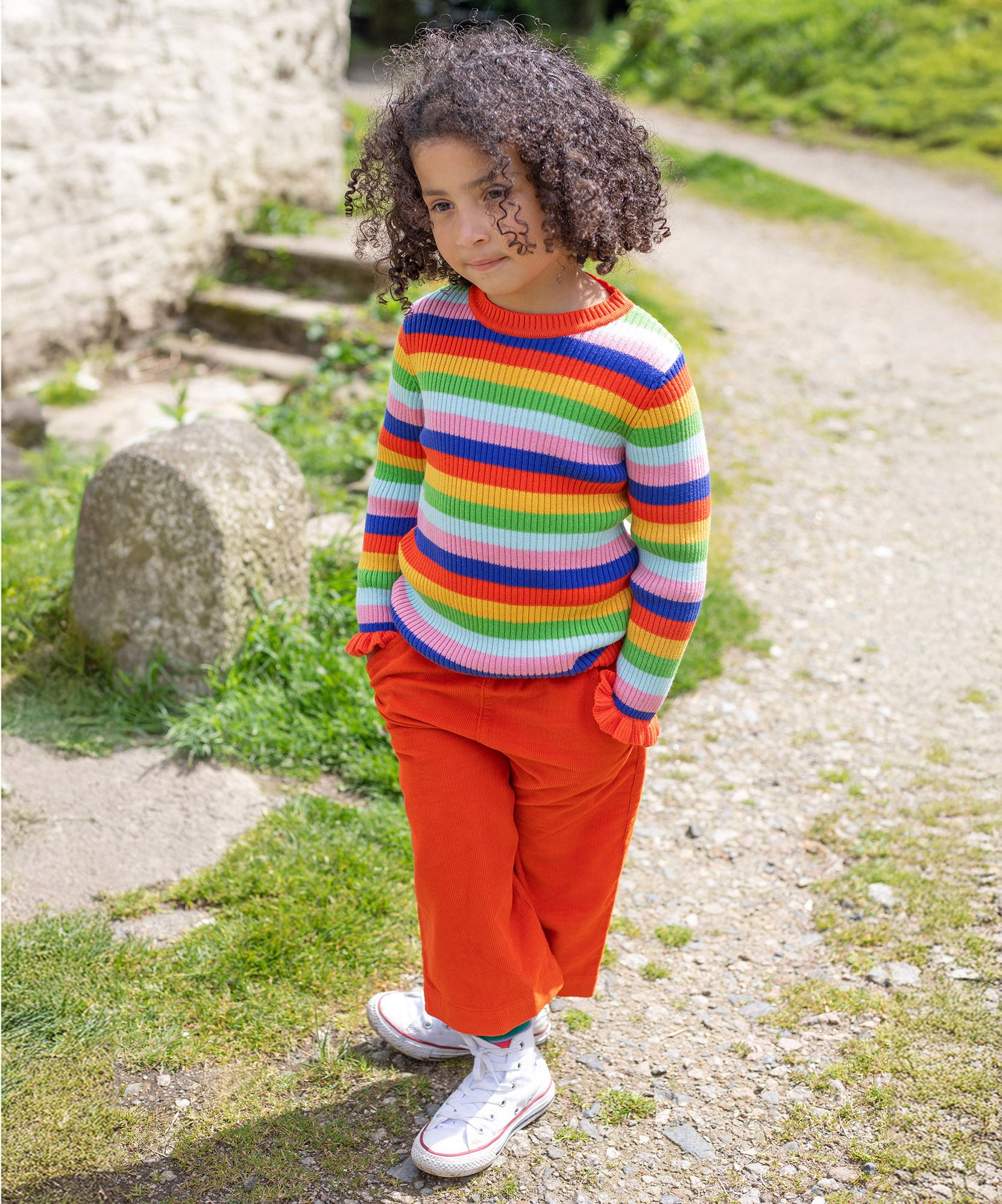 A child is stood outside in the sunshine, with their hands in their pockets. The child wears the Frugi Zoe Knitted Jumper - Retro Stripe, which looks very vibrant in colour in the natural day light