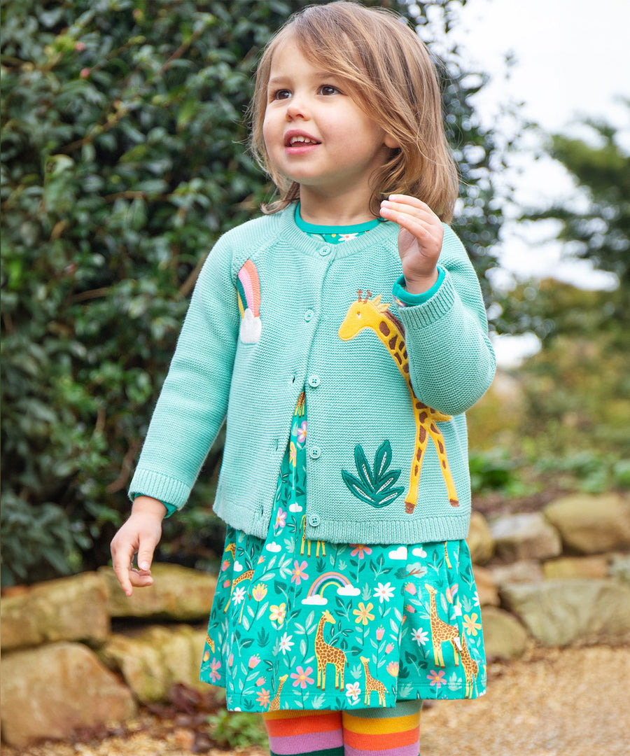 A child outside, wearing the Frugi Colby Cardigan - Moss/Giraffe and a Frugi giraffe dress