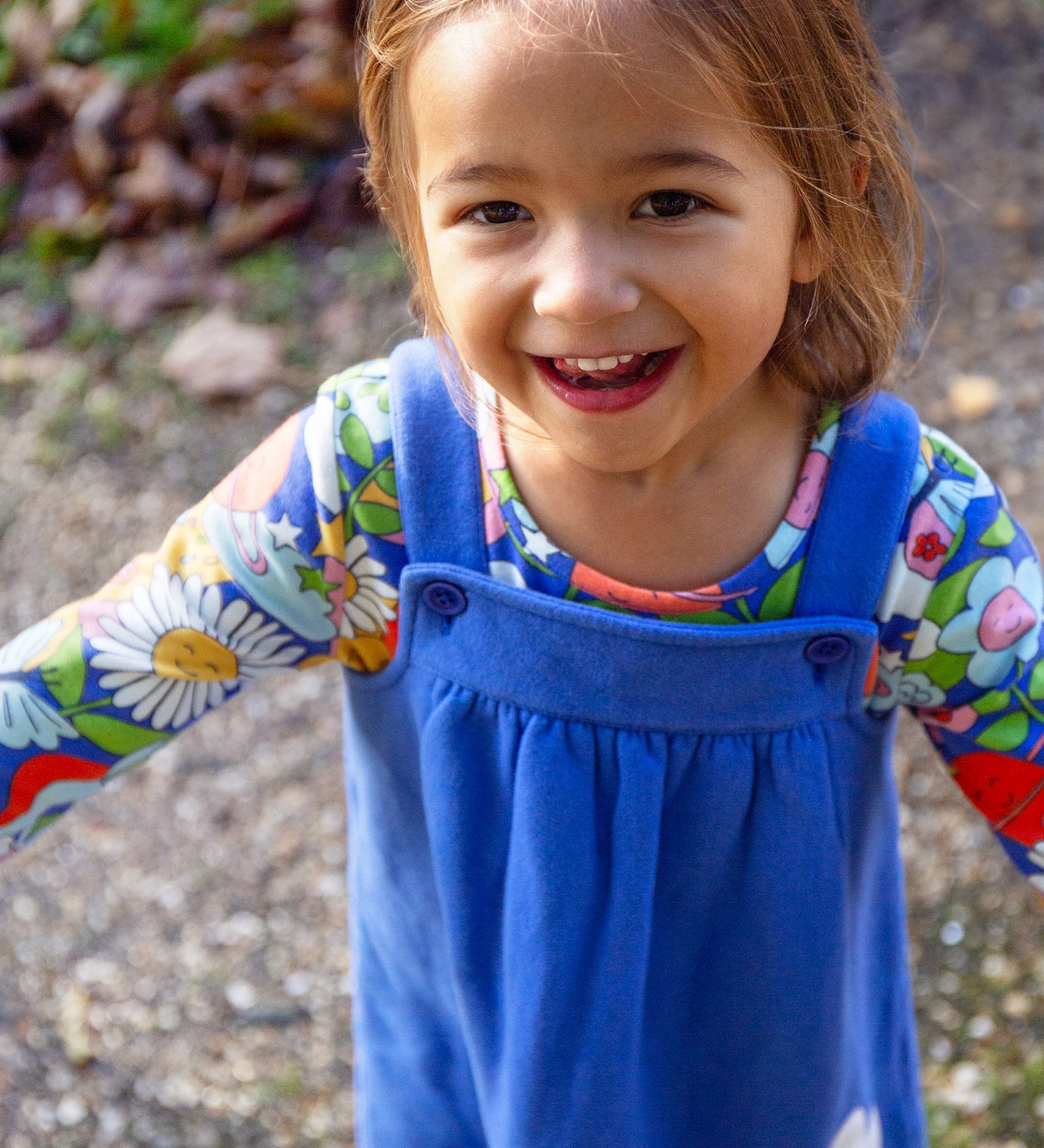 A child outside in the sun, smiling and with their arms out stretched to the side. The child is wearing the Frugi Polly Pinafore Outfit - Blue Tang/Retro Happy. The image showing the adjustable straps with adjust using a button