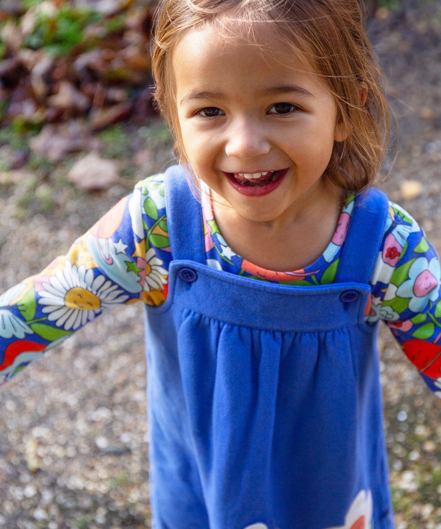 A child outside in the sun, smiling and with their arms out stretched to the side. The child is wearing the Frugi Polly Pinafore Outfit - Blue Tang/Retro Happy. The image showing the adjustable straps with adjust using a button