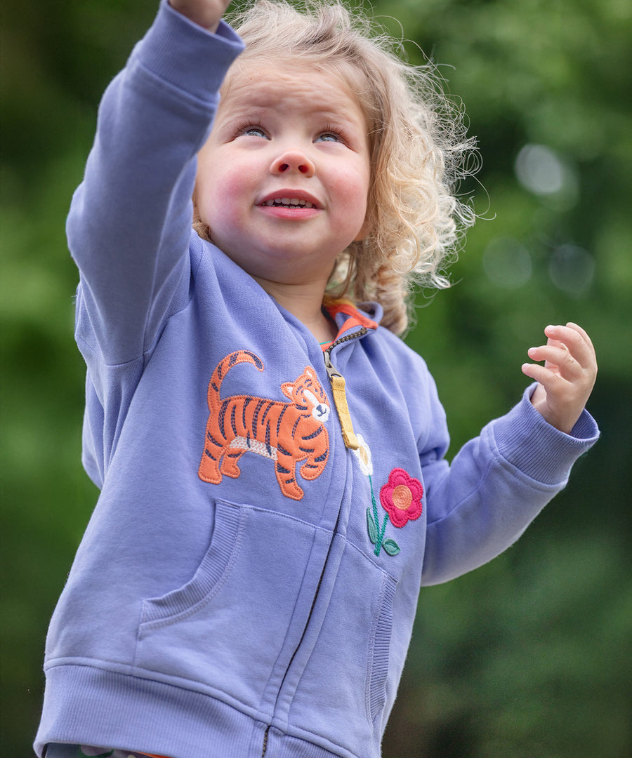 A child playing outside in front of green trees wearing the Frugi Carbis Hoodie - Grape/Tiger