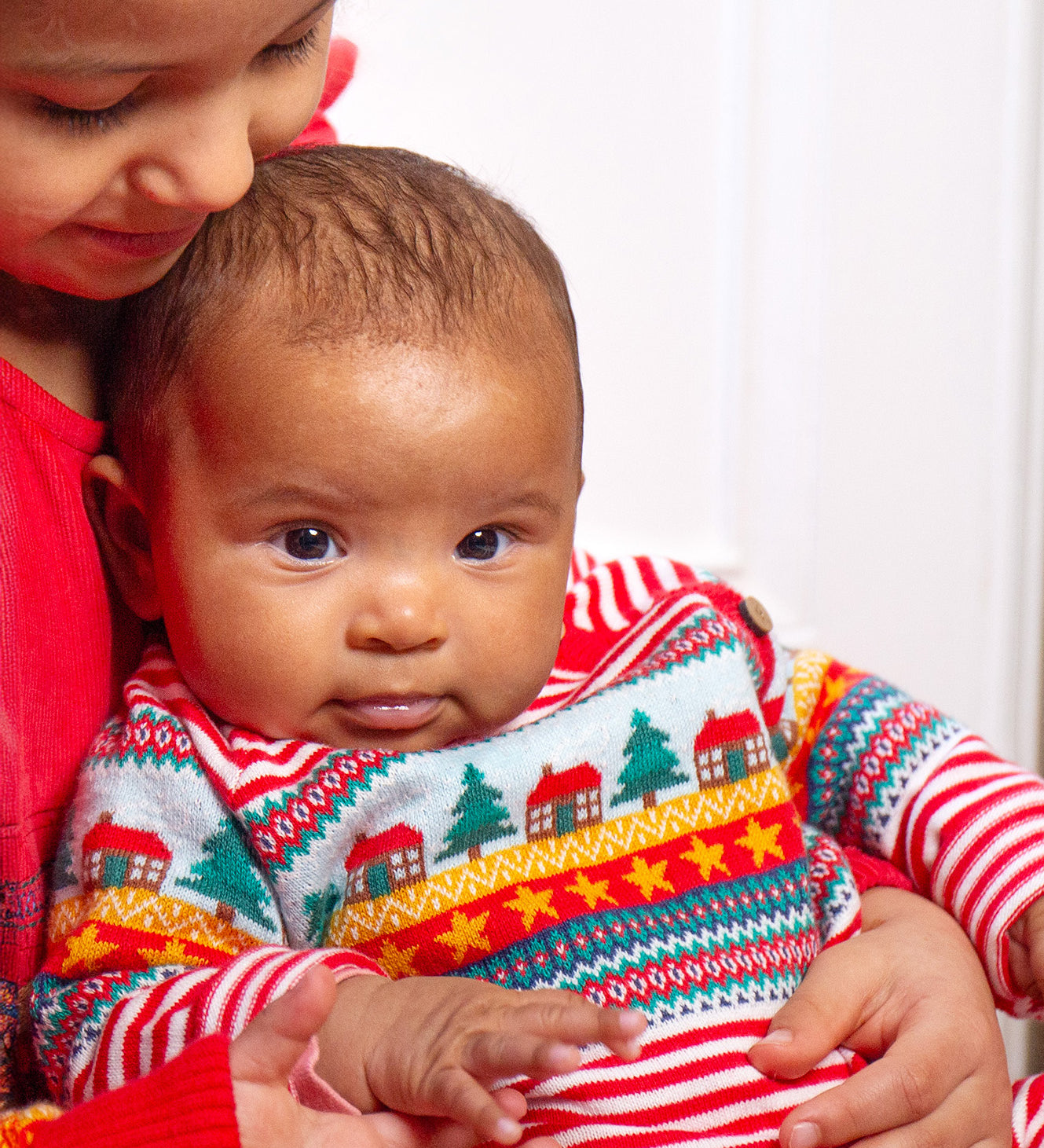 A child looking forwards wearing the Frugi Lumi Knitted Romper - Fairisle Christmas Stripe, sitting with another child