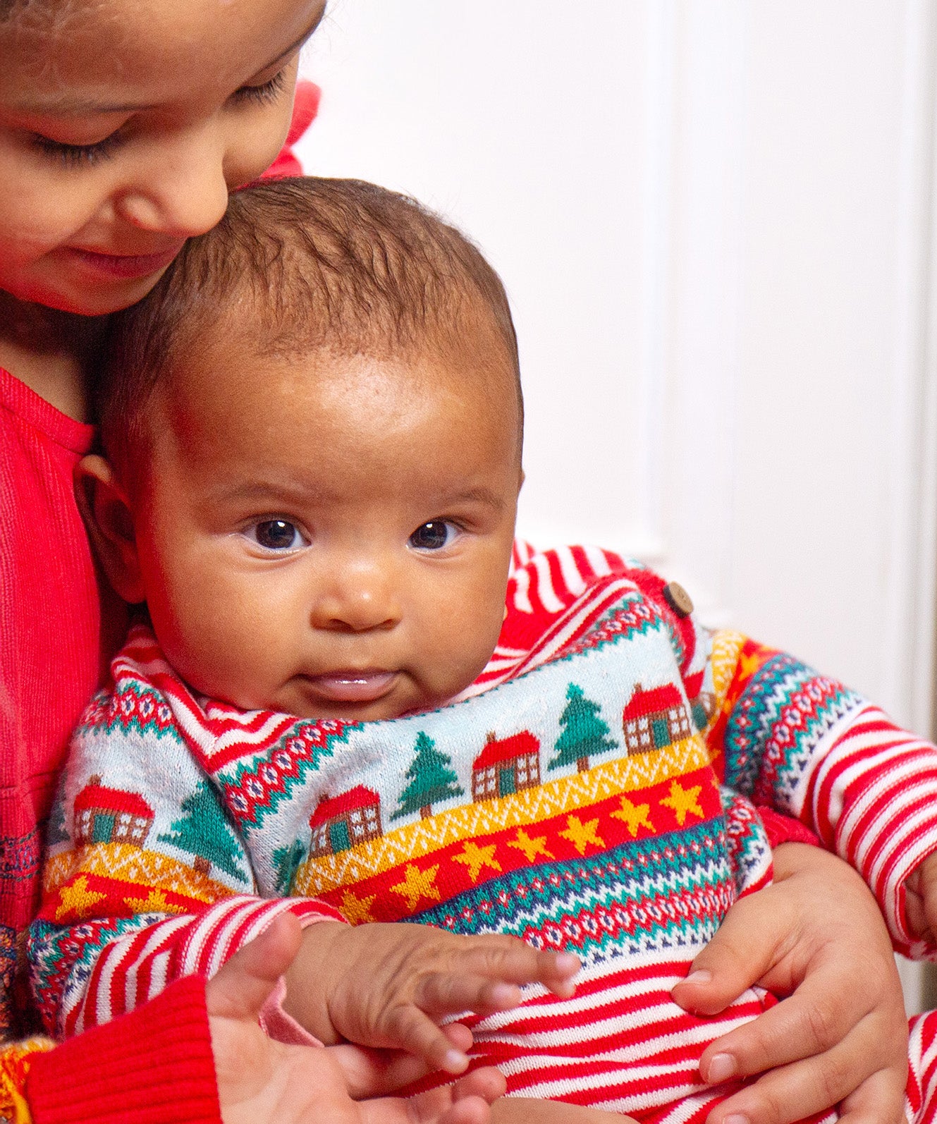 A child looking forwards wearing the Frugi Lumi Knitted Romper - Fairisle Christmas Stripe, sitting with another child