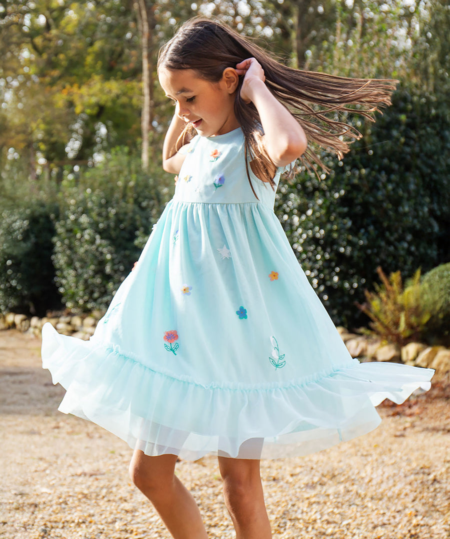 A child is wearing the Frugi Tilda Tulle Dress - Glacia/Floral outside in the sunshine. The child is twirling around in the dress, to show the ruffles and and how well the fabric flows.