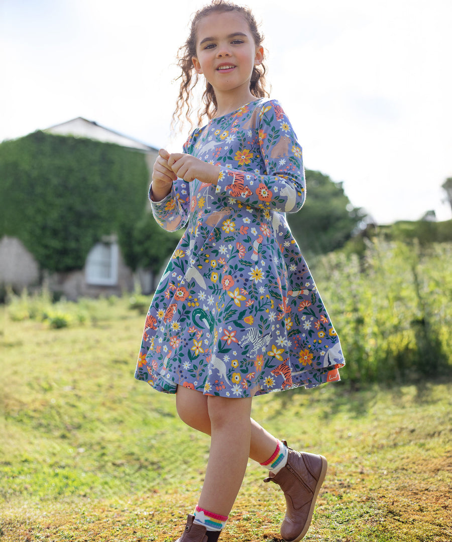 A child happily outside in a grassy green field and a building in the background, wearing the Frugi Sofia Skater Dress - Rainforest Friends