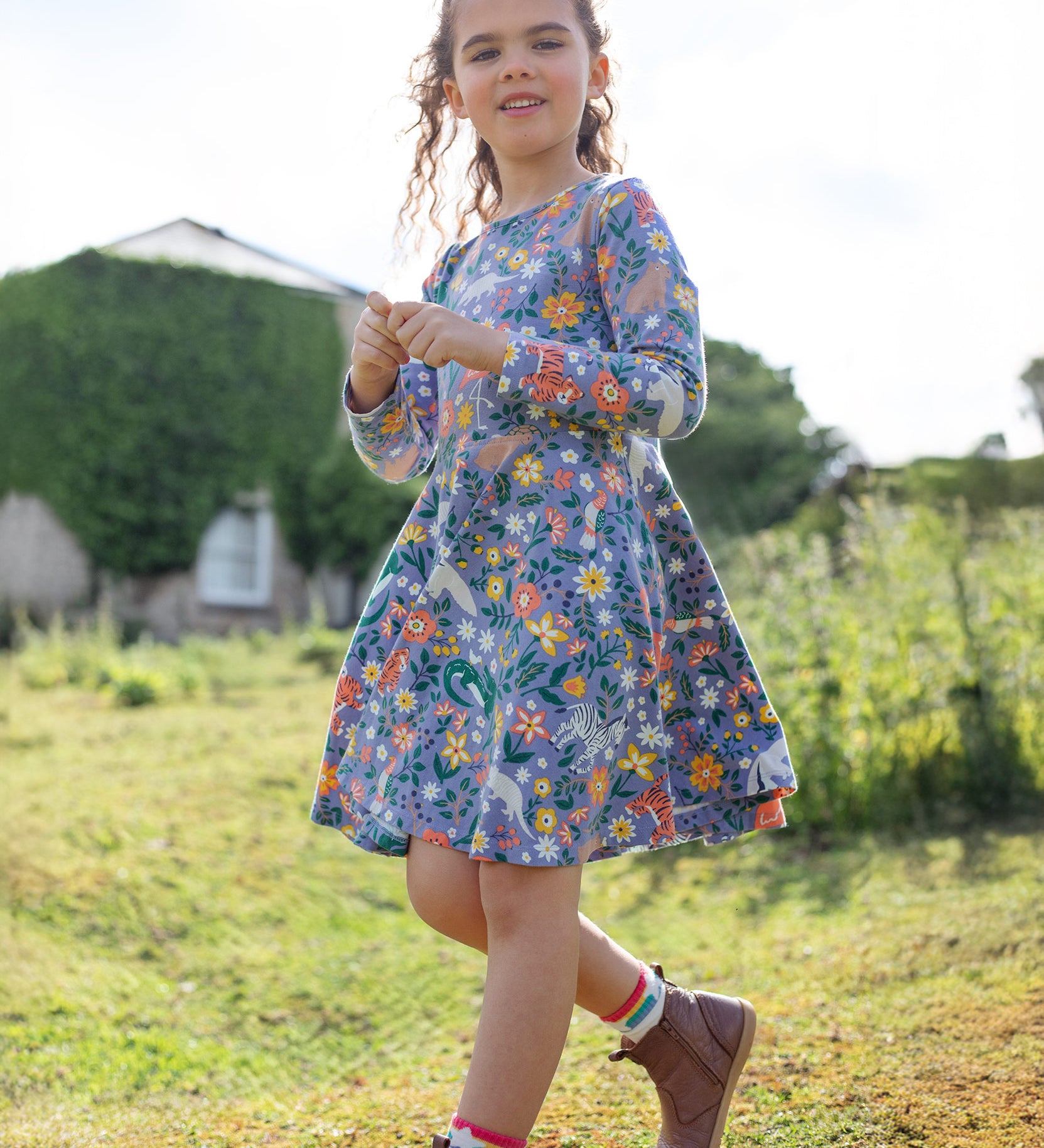 A child happily outside in a grassy green field and a building in the background, wearing the Frugi Sofia Skater Dress - Rainforest Friends