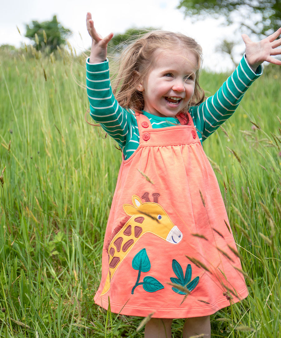 A child playing outside in a grassy field, wearing the Frugi Paige Pinafore Outfit - Cantaloupe/Iguana Breton