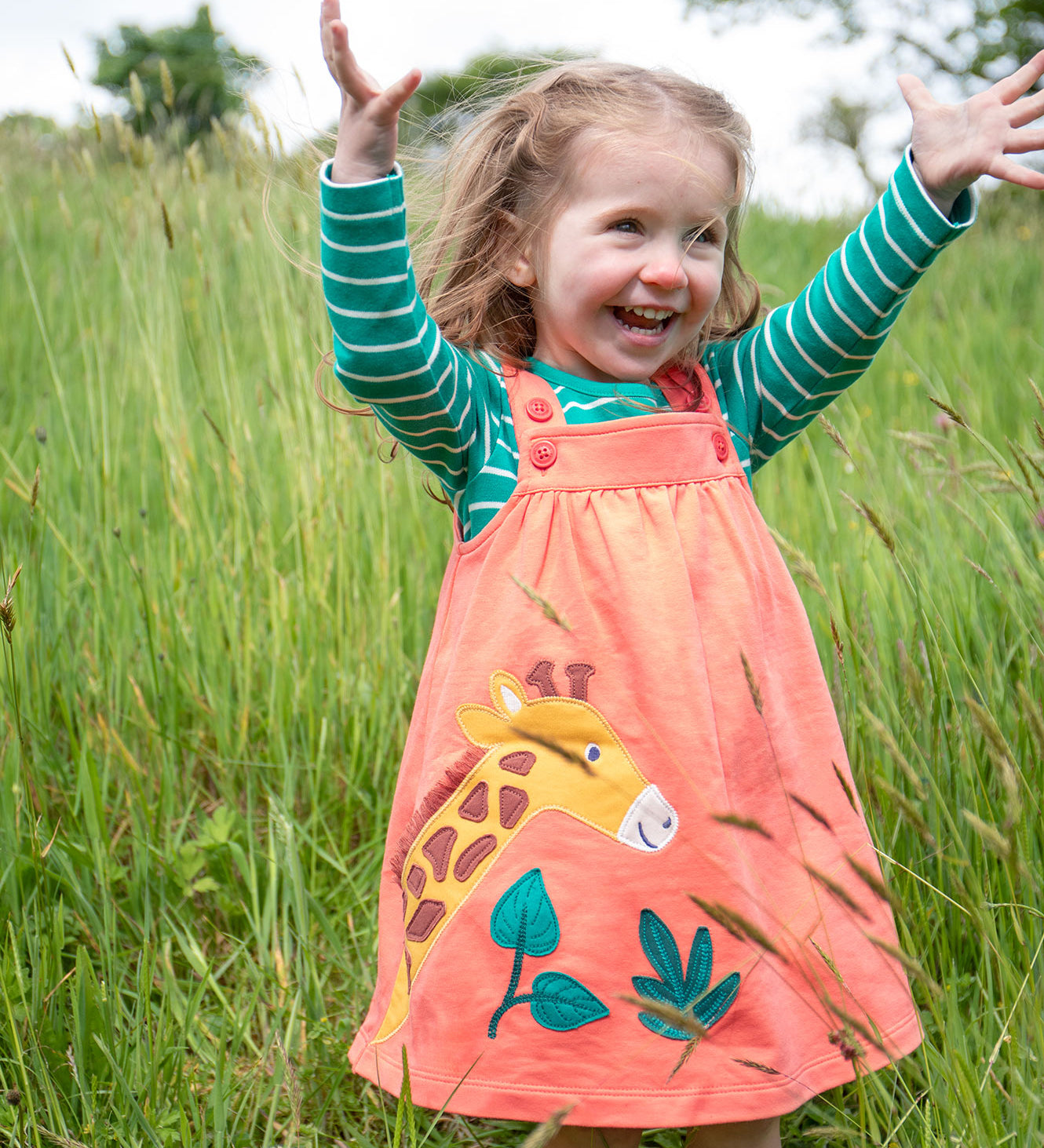 A child playing outside in a grassy field, wearing the Frugi Paige Pinafore Outfit - Cantaloupe/Iguana Breton