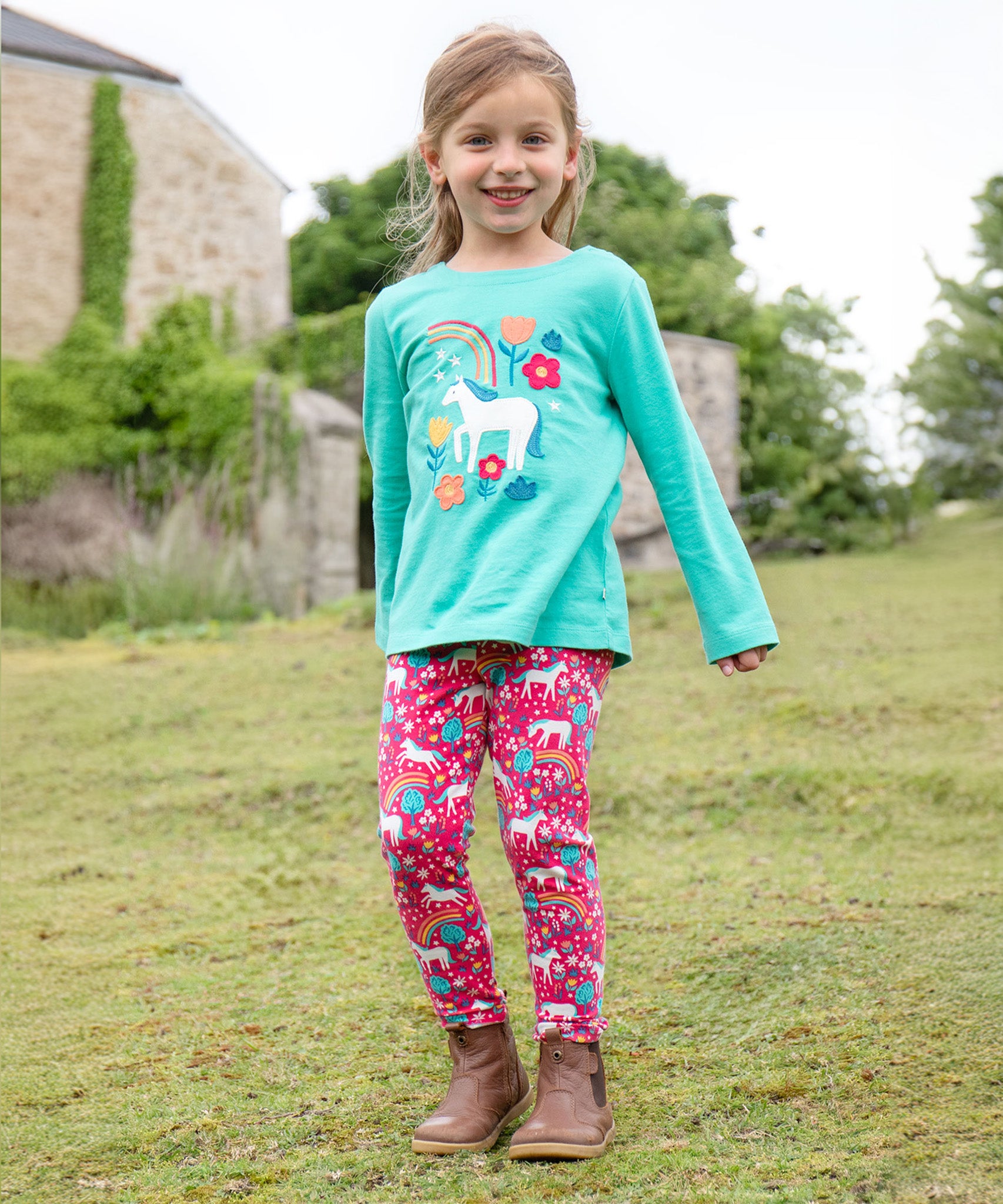 A child outside wearing the Frugi Anna Applique Top - Pacific Aqua/Horse, and Frugi Wild Horses leggings