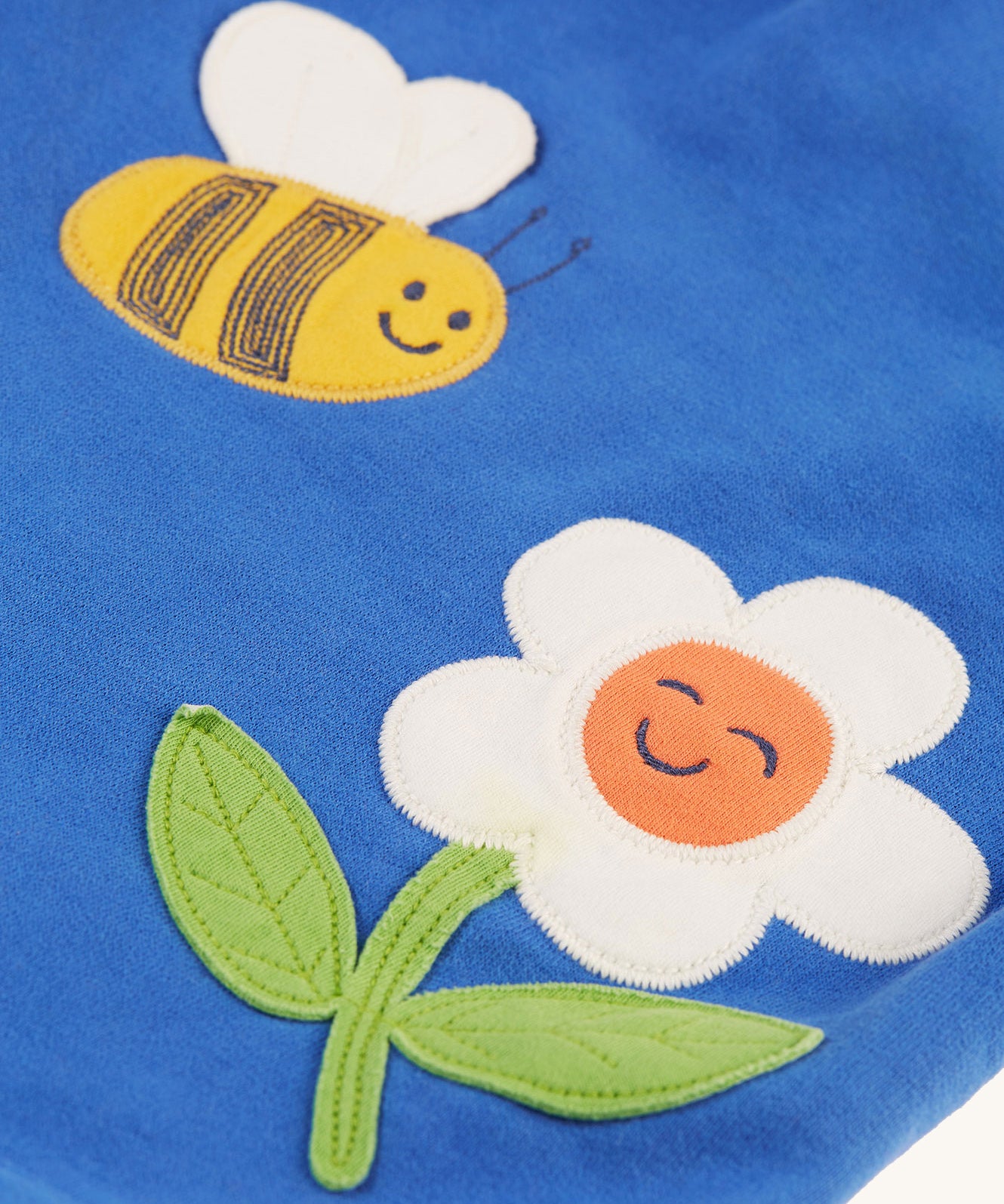 A closer look at the bee and flower applique on the Frugi Polly Pinafore Outfit - Blue Tang/Retro Happy. The appliques' also include some embroidery detail like the black stripes on the bee, and the leaf vein pattern on the flower. The applique's are on the blue fabric of the dress