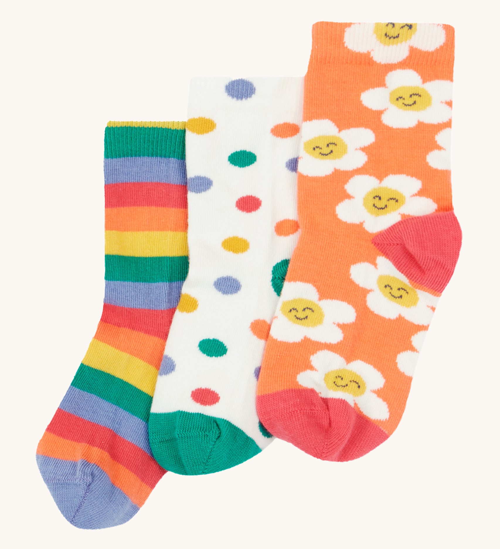 Frugi Rock My Socks - Stay Wild 3 Pack. 3 pairs of soft, organic cotton, kids socks in the Stay Wild prints. from left to right, the left sock is a rainbow stripe print in lilac, orange, red, green and yellow. The middle sock is white with colourful polka dots and a green toe and heel, and the last sock is orange with white, smiling flowers and a red toe and heel.