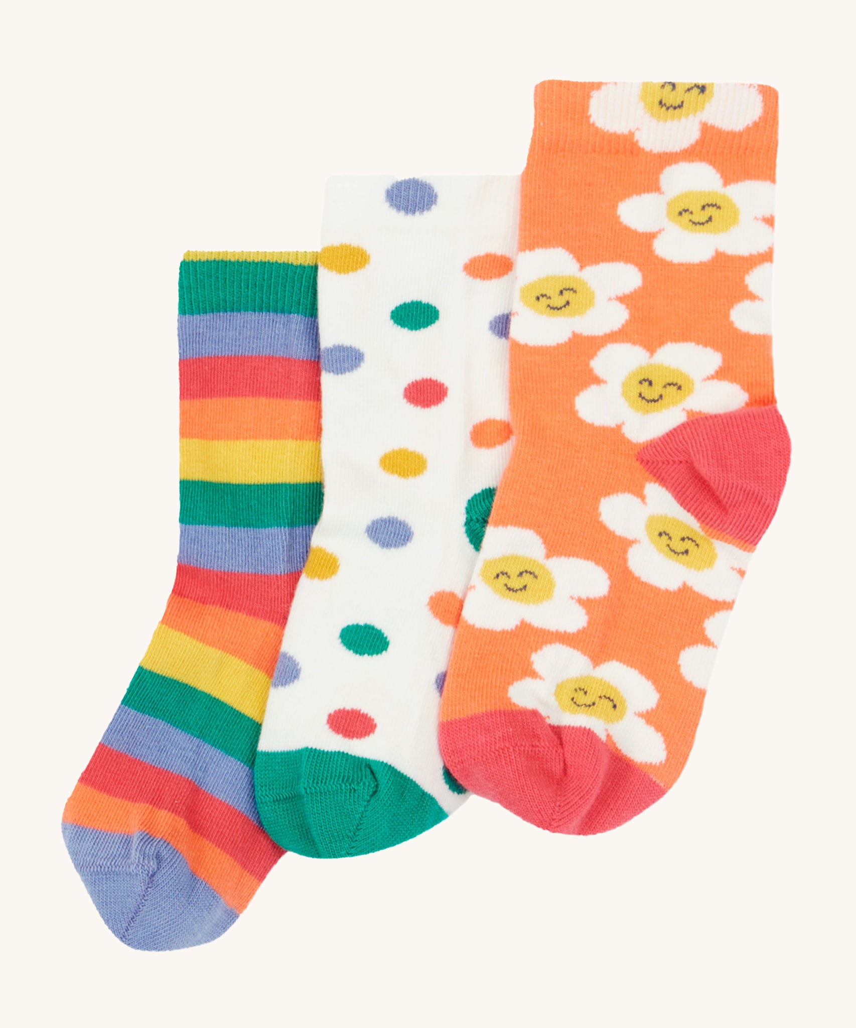 Frugi Rock My Socks - Stay Wild 3 Pack. 3 pairs of soft, organic cotton, kids socks in the Stay Wild prints. from left to right, the left sock is a rainbow stripe print in lilac, orange, red, green and yellow. The middle sock is white with colourful polka dots and a green toe and heel, and the last sock is orange with white, smiling flowers and a red toe and heel.