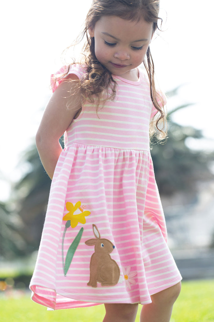 A young girl wearing a pink and white striped Frugi dress with adorable appliqué details featuring a brown bunny, a daffodil, and a flower. The soft organic cotton and playful design capture Frugi’s signature style—bright, comfortable, and perfect for little adventures. Showcasing the Frugi sale at Babipur