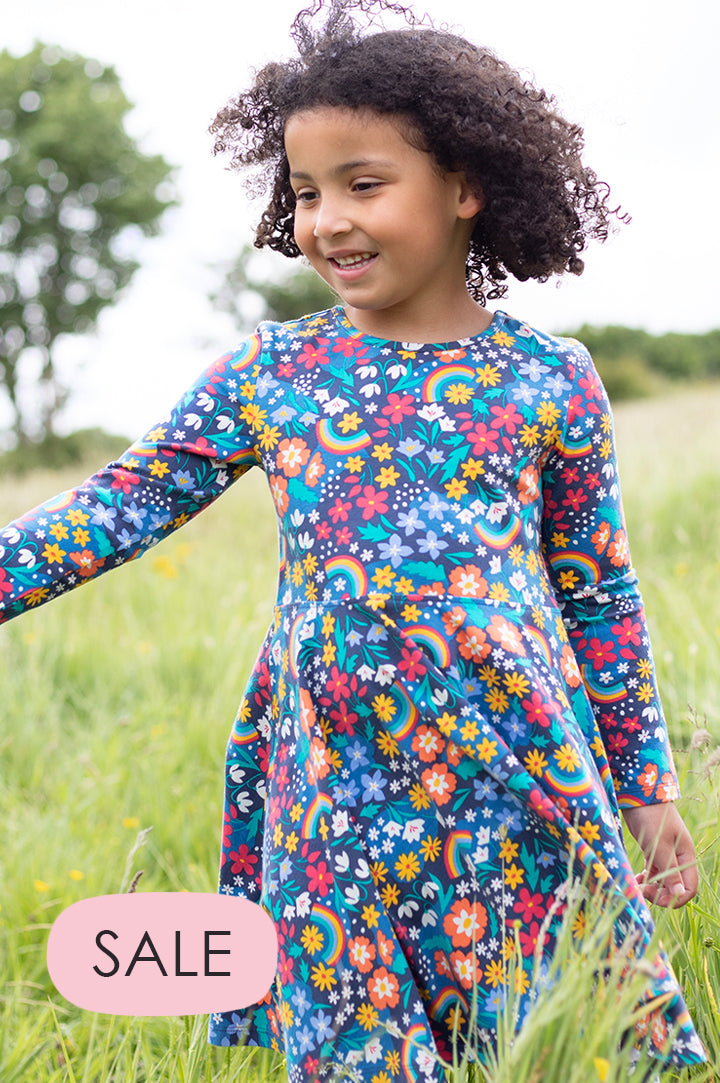 A young girl with curly hair walking through a field wearing Frugi Sofia Skater Dress Winter Hedgerow. GOTS organic dark blue long sleeve dress with a busy and colourful print featuring rainbows and winter flowers.