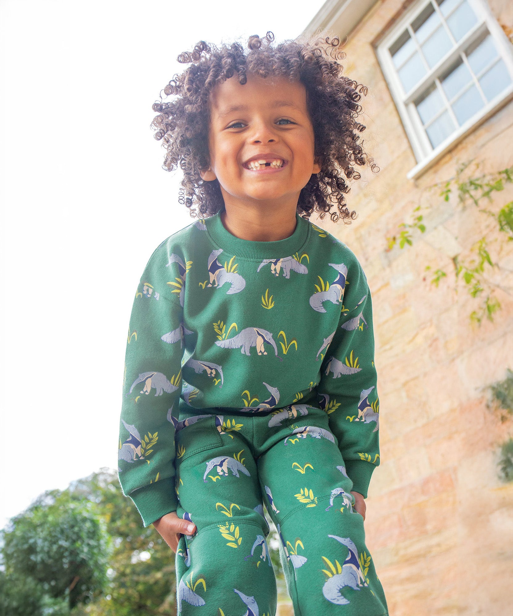 A child happily playing outside next to a house, wearing the Frugi Superb Sweatshirt - Anteater Parade and Frugi Malpas Joggers - Anteater Parade