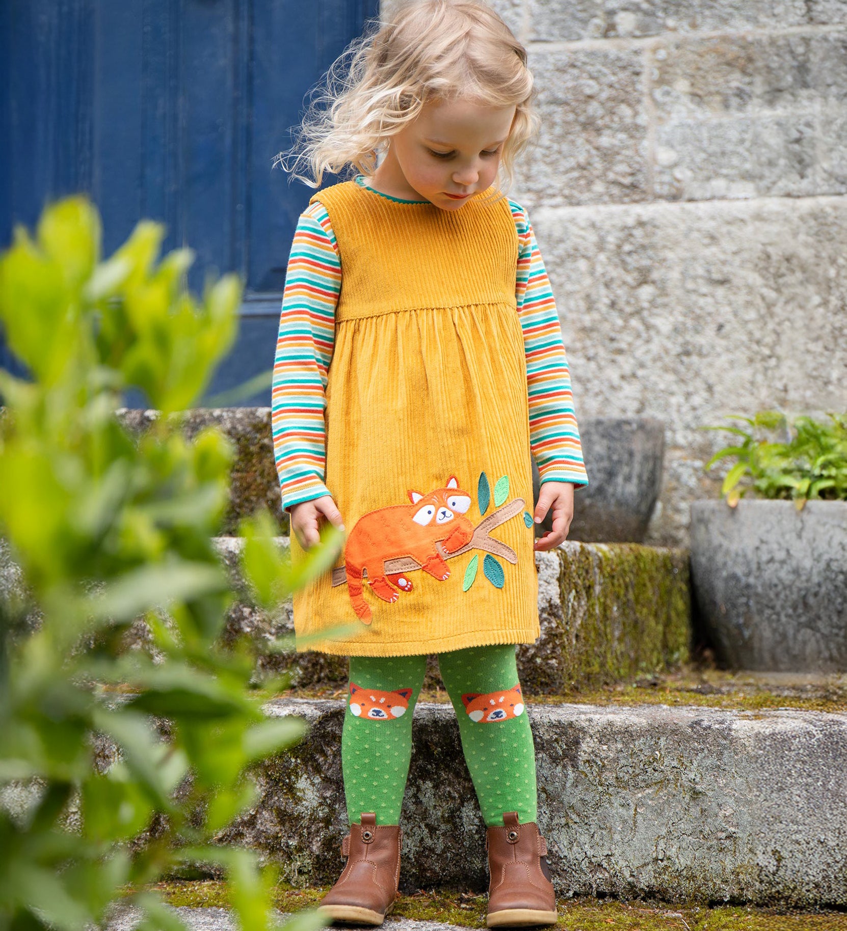 A child stood outside a building and looking down. The child is wearing the Frugi Lovely Lily Cord Dress - Gold/Red Panda, and the Frugi Norah Tights - Red Panda Pals, along with a Frugi rainbow stripe top underneath the dress.