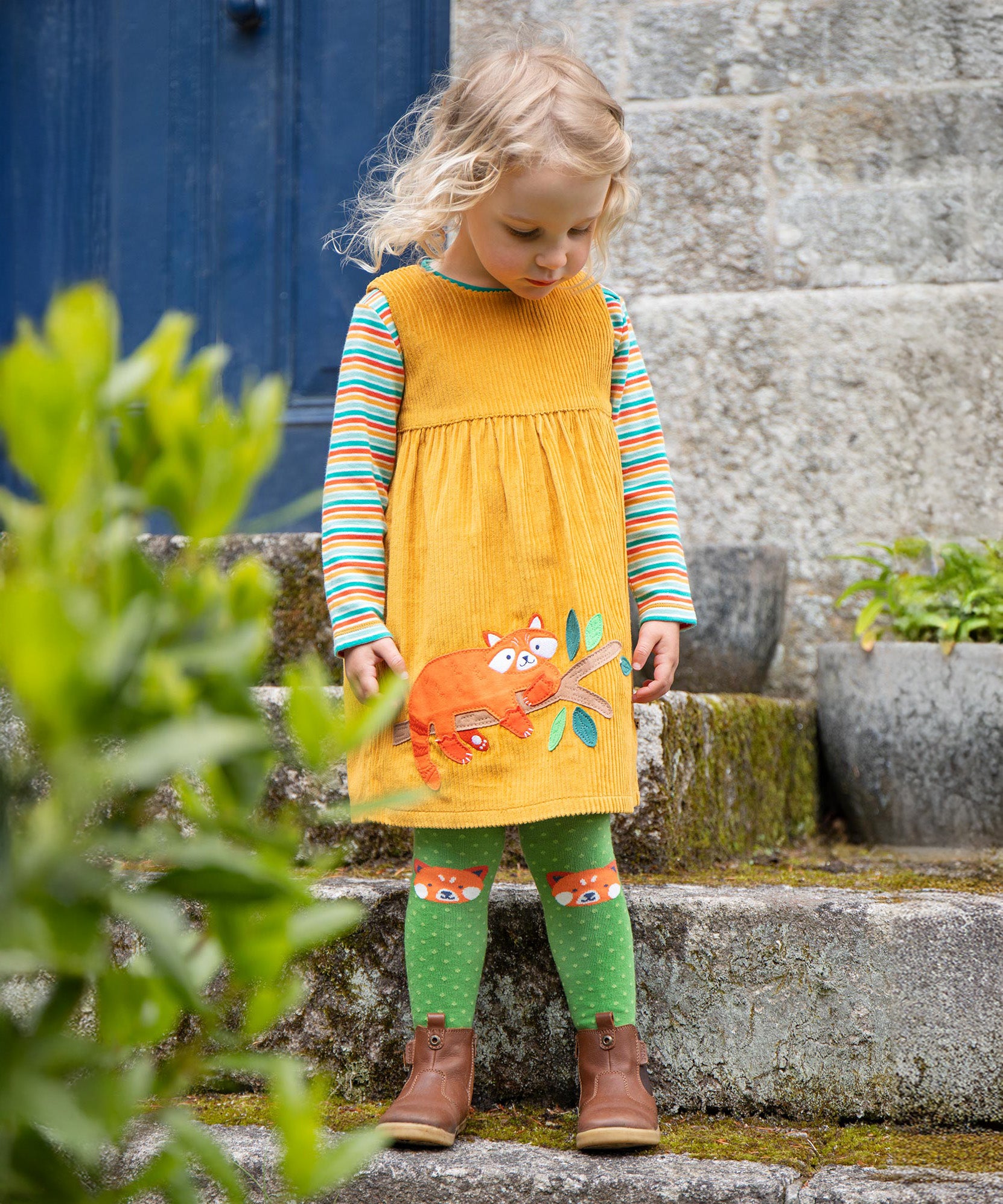 A child stood outside a building and looking down. The child is wearing the Frugi Lovely Lily Cord Dress - Gold/Red Panda, and the Frugi Norah Tights - Red Panda Pals, along with a Frugi rainbow stripe top underneath the dress.