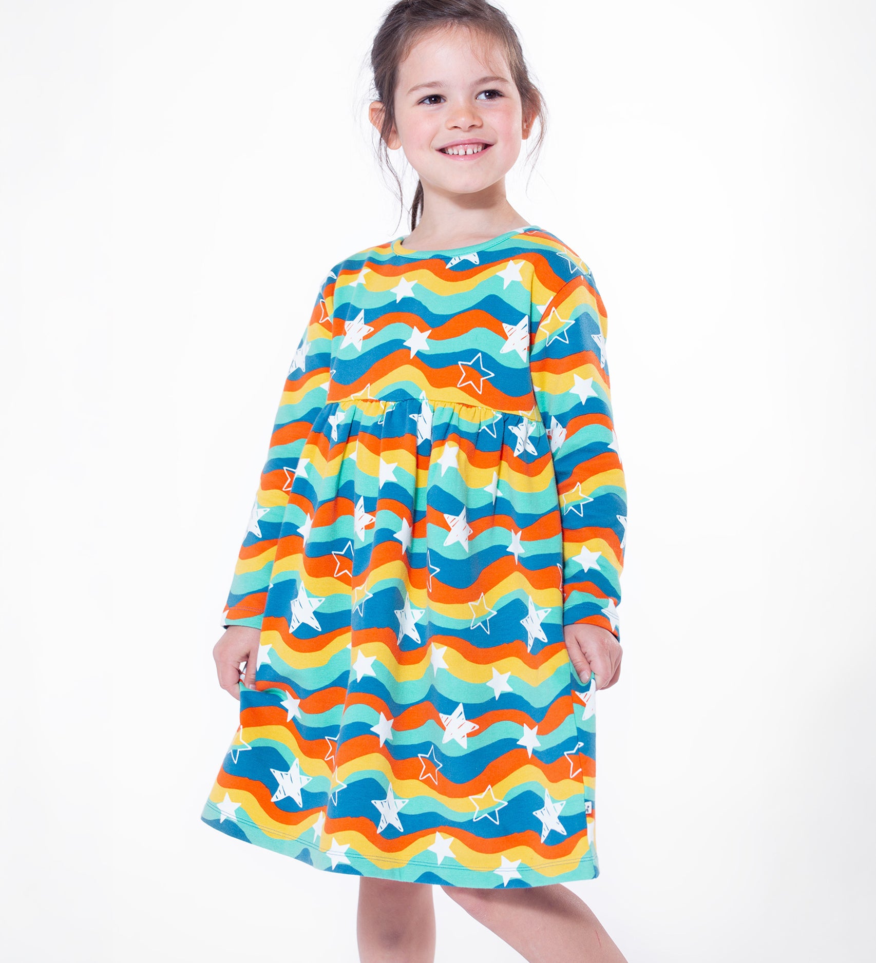 A child stood on a white background, wearing the Frugi Cosy Amelia Dress - Wavy Stars