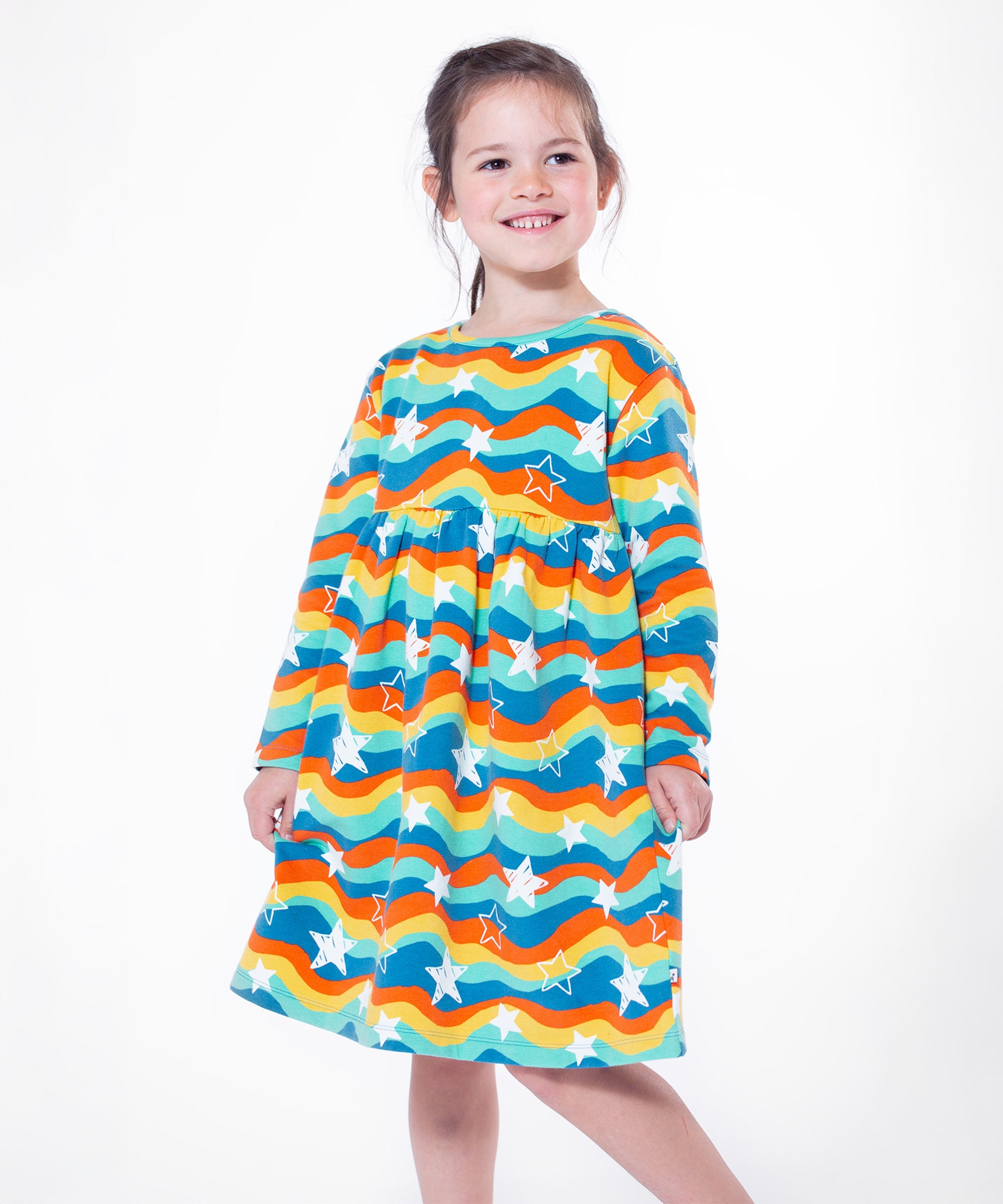 A child stood on a white background, wearing the Frugi Cosy Amelia Dress - Wavy Stars