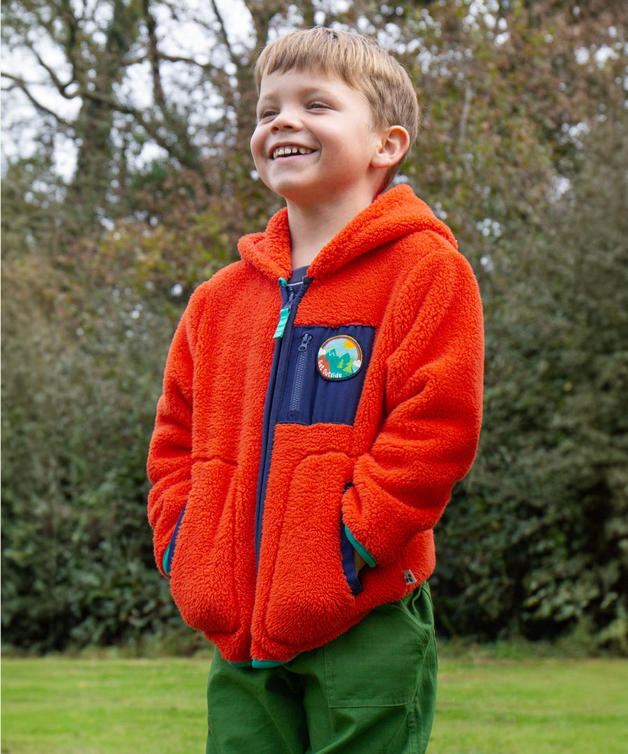 A child outside wearing the Frugi Toby Ted Fleece Jacket - Bonfire, with green Frugi trousers